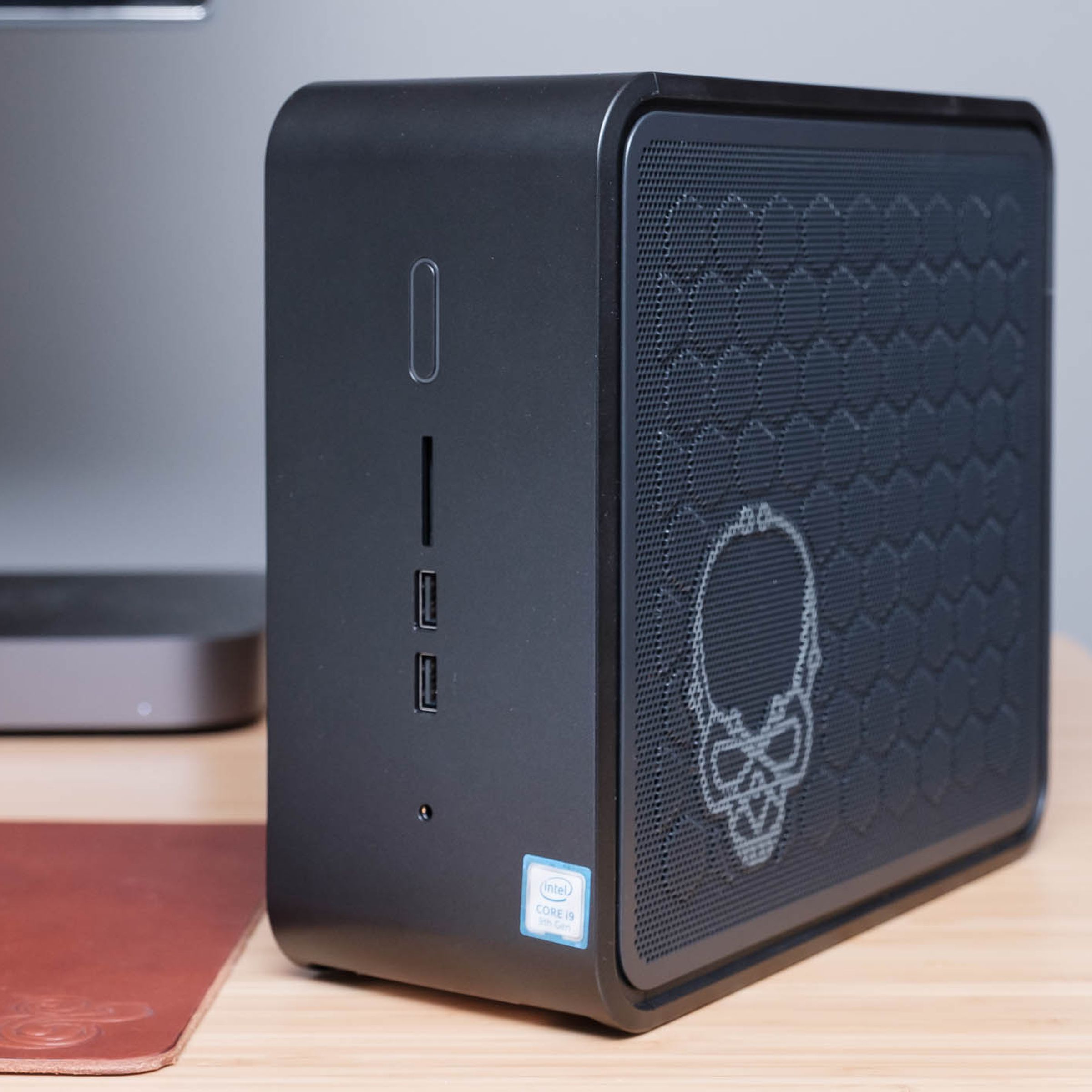 Intel NUC 9 computer standing vertically on a wood desk with a skull etched on the side and a gray Mac Mini in the background