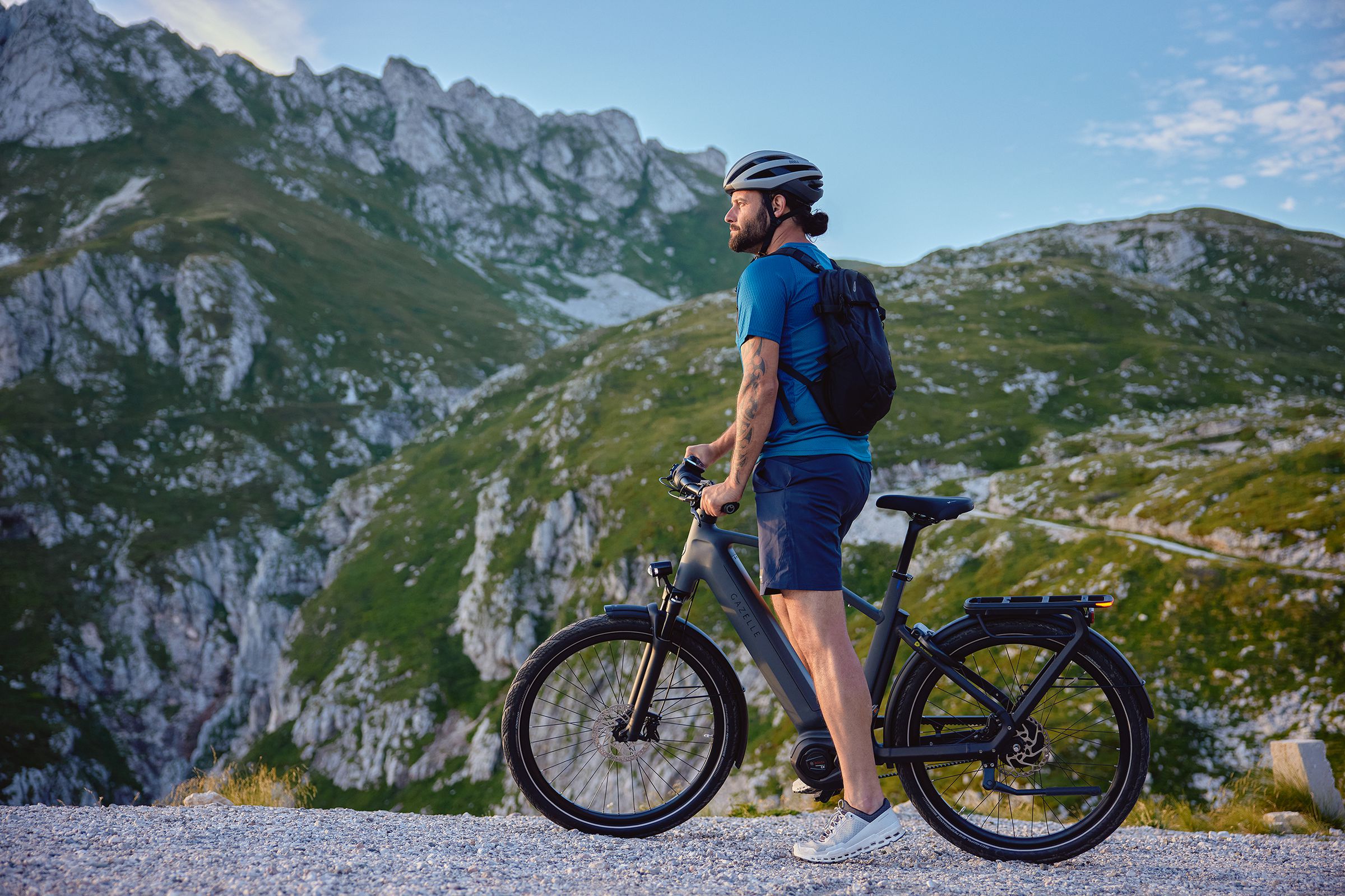 A man straddles his Gazelle e-bike while looking down the road ahead as a snow-capped mountain cap looms in the distance.