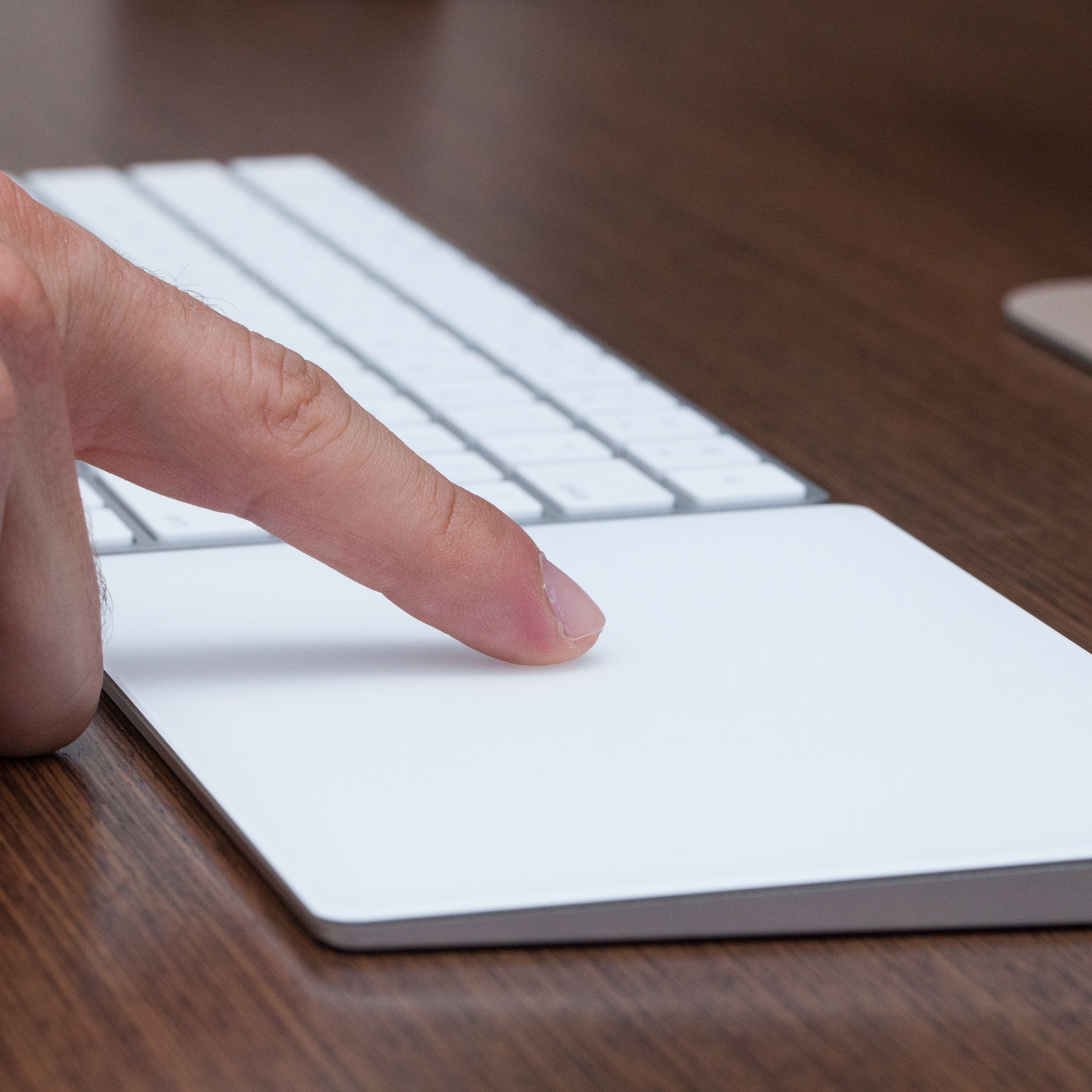 A finger touching Apple’s Magic Trackpad, which sits next to a keyboard on a desk.