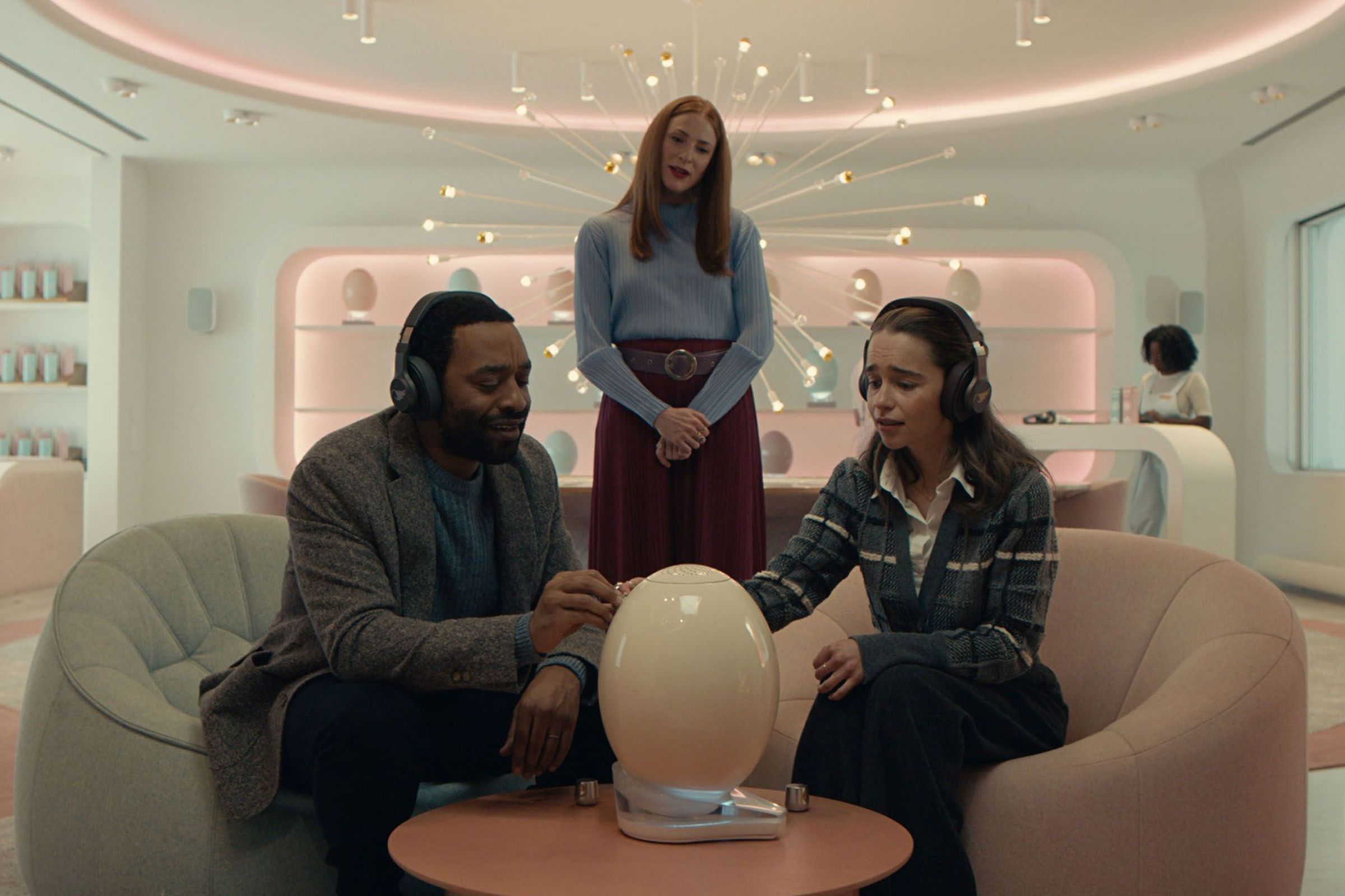 A man and a woman wearing headphones and sitting on separate loveseats as they stroke and awe at a massive, gleaming egg placed on a table between them. Behind the couple is a woman gazing down at them both while she tells them about what they’re hearing from the egg. Around them all you can see that they’re in some sort of luxury showroom for the strange eggs, more of which line the wall in holsters in the distance.