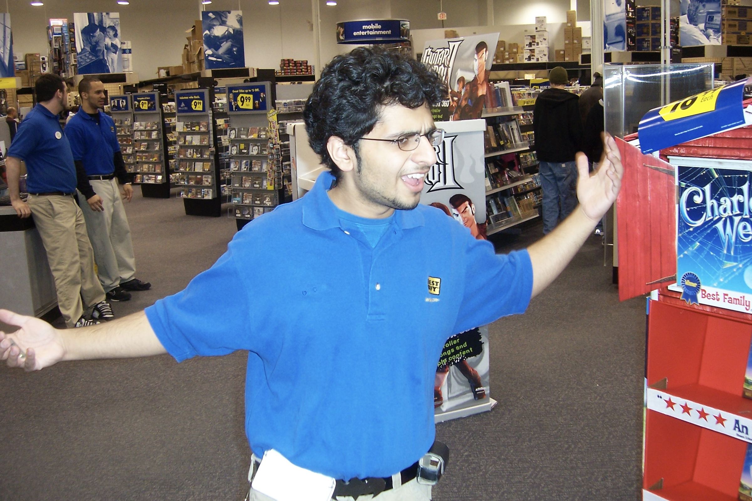 Best Buy employee in blue shirt standing in front of a Guitar Hero 2 stand and movie and CD aisles in the back, employee waving arms