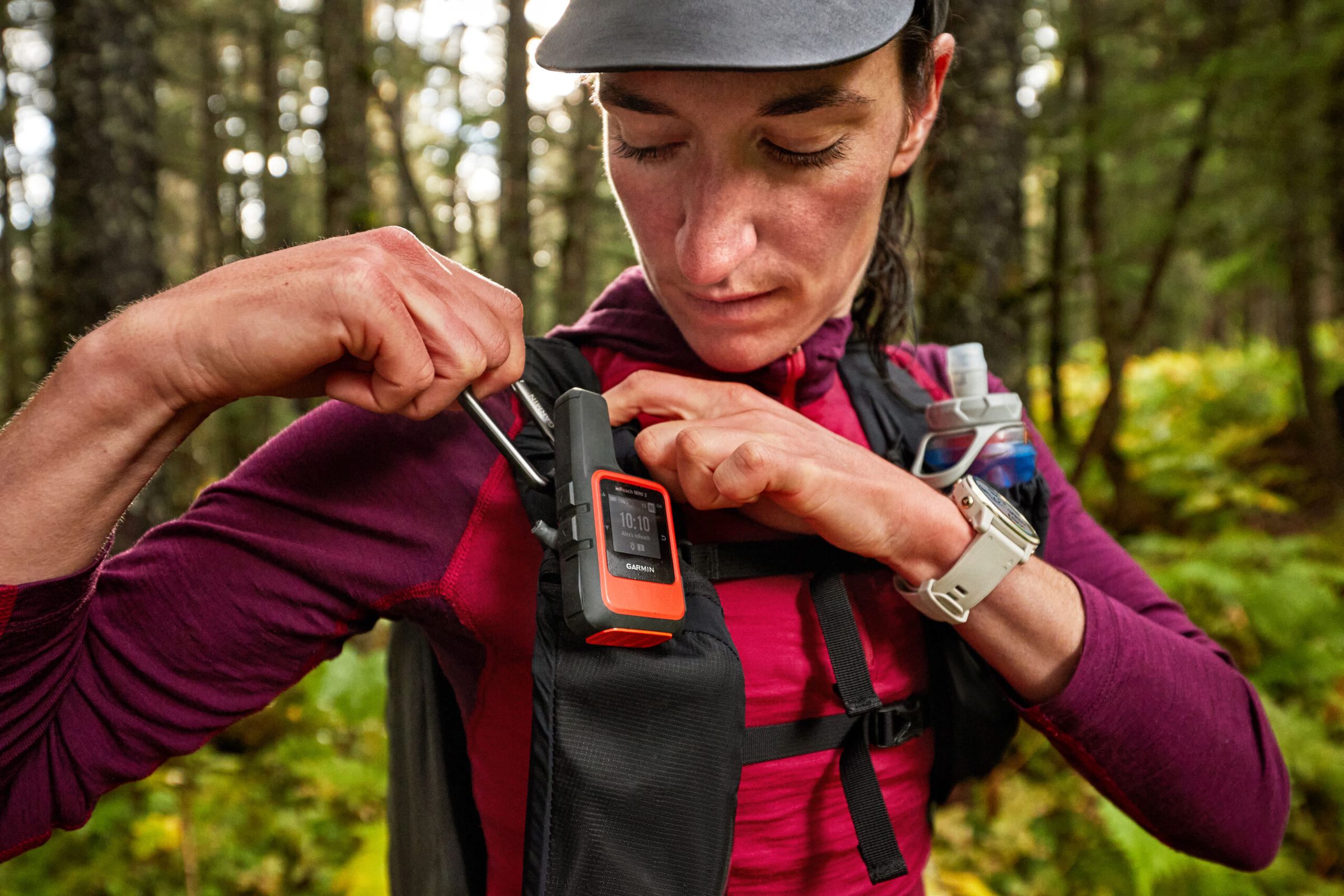 An image of woman in the woods clipping a Garmin InReach Mini 2 to the right shoulder strap of her backpack.