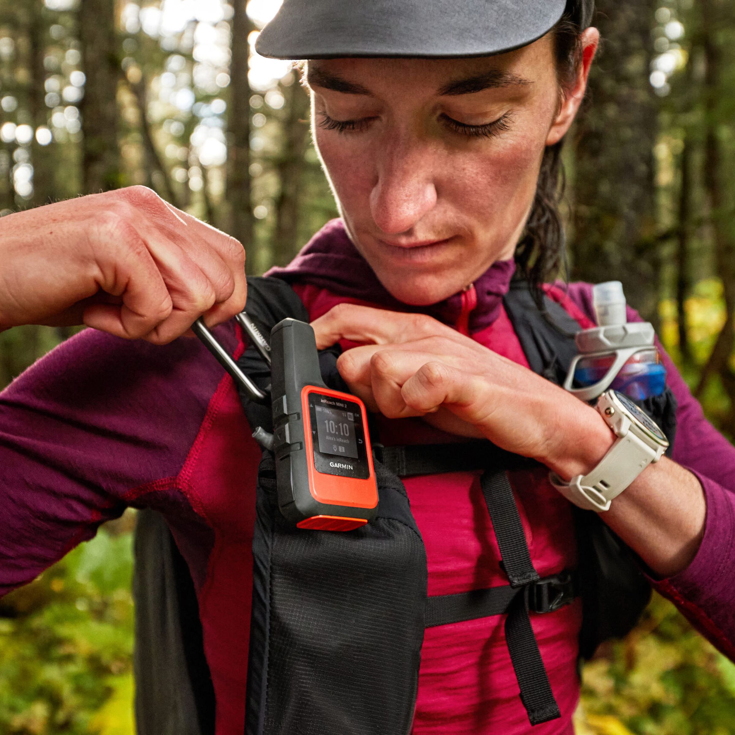 An image of woman in the woods clipping a Garmin InReach Mini 2 to the right shoulder strap of her backpack.