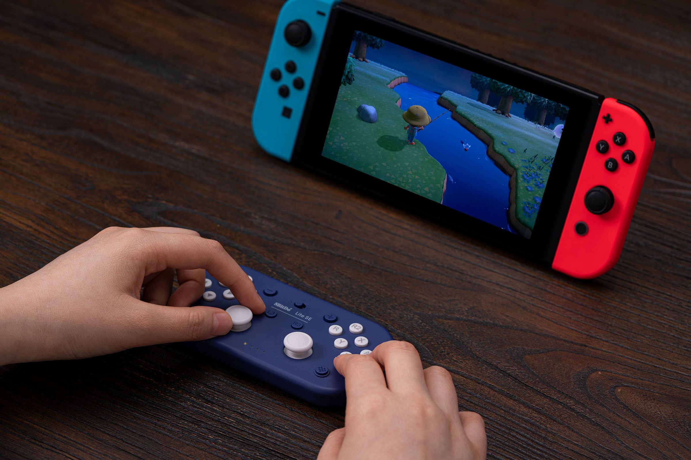 The 8BitDo controller being used on a tabletop. The player’s hands are visible, with the left controlling the left analog stick and the right on the A and B buttons. A Nintendo Switch in stand mode is in the background.