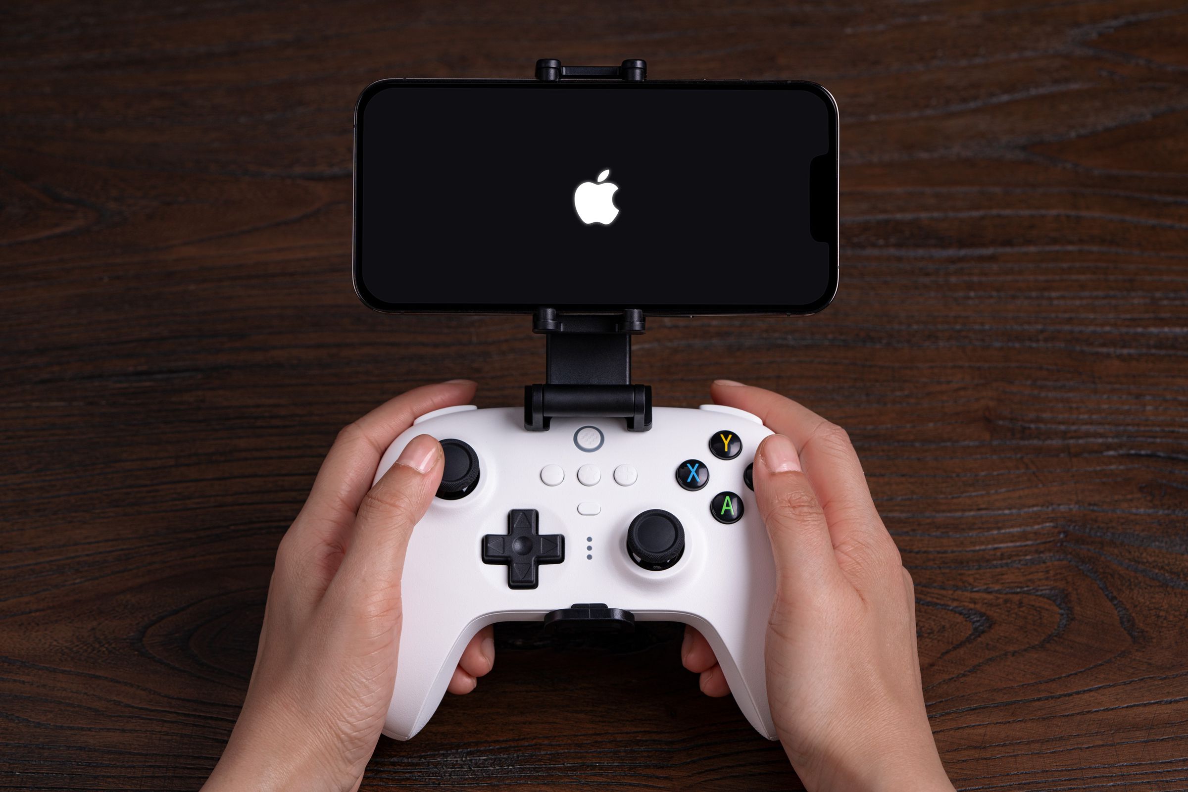 An 8BitDo wireless game controller with an Apple iPhone attached to it via a mounting clip, held by a pair of hands.