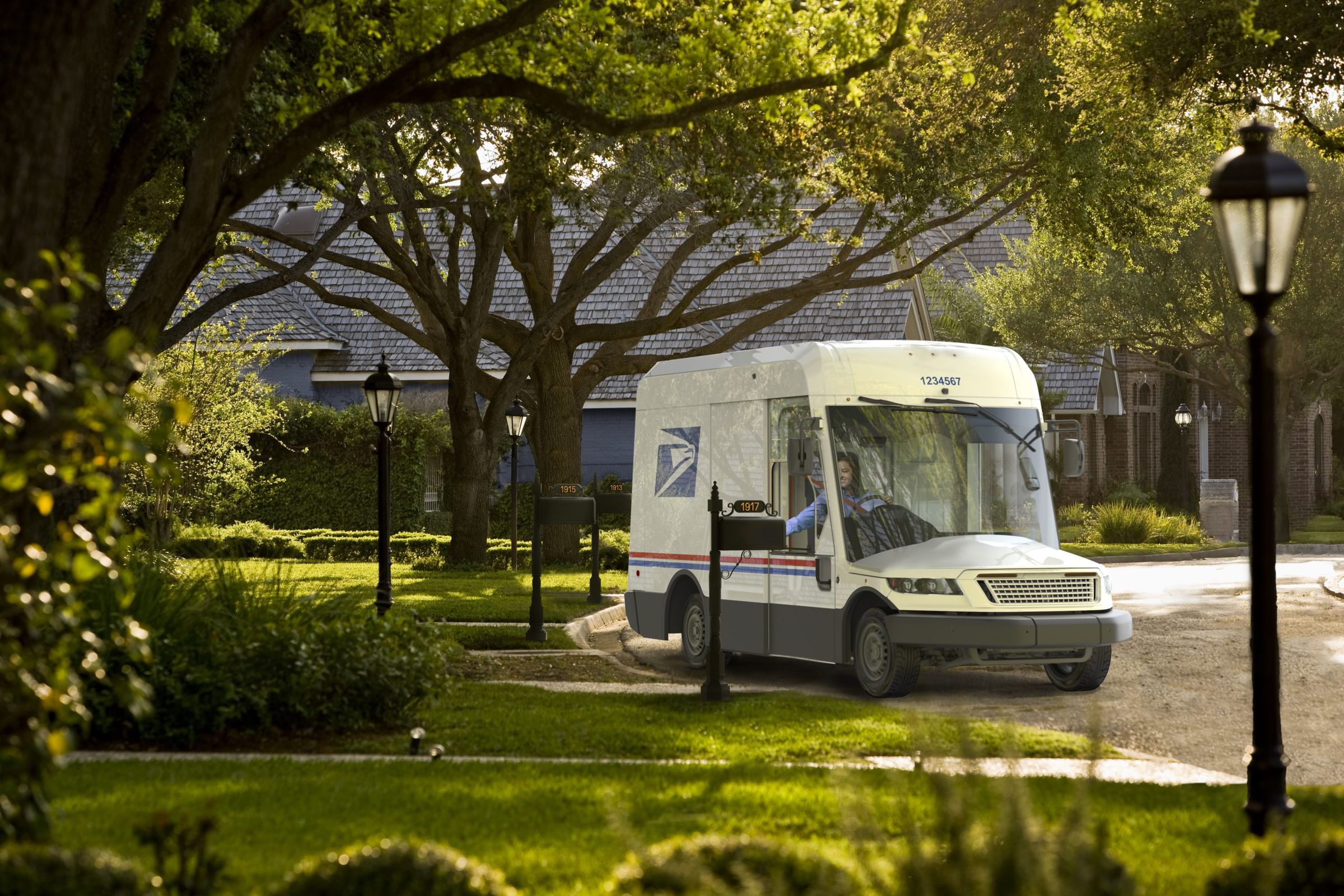 In a lush green suburban neighborhood, a friendly USPS white truck with a new design that has a huge windshield and low to the ground hood is by a driveway delivering letters.