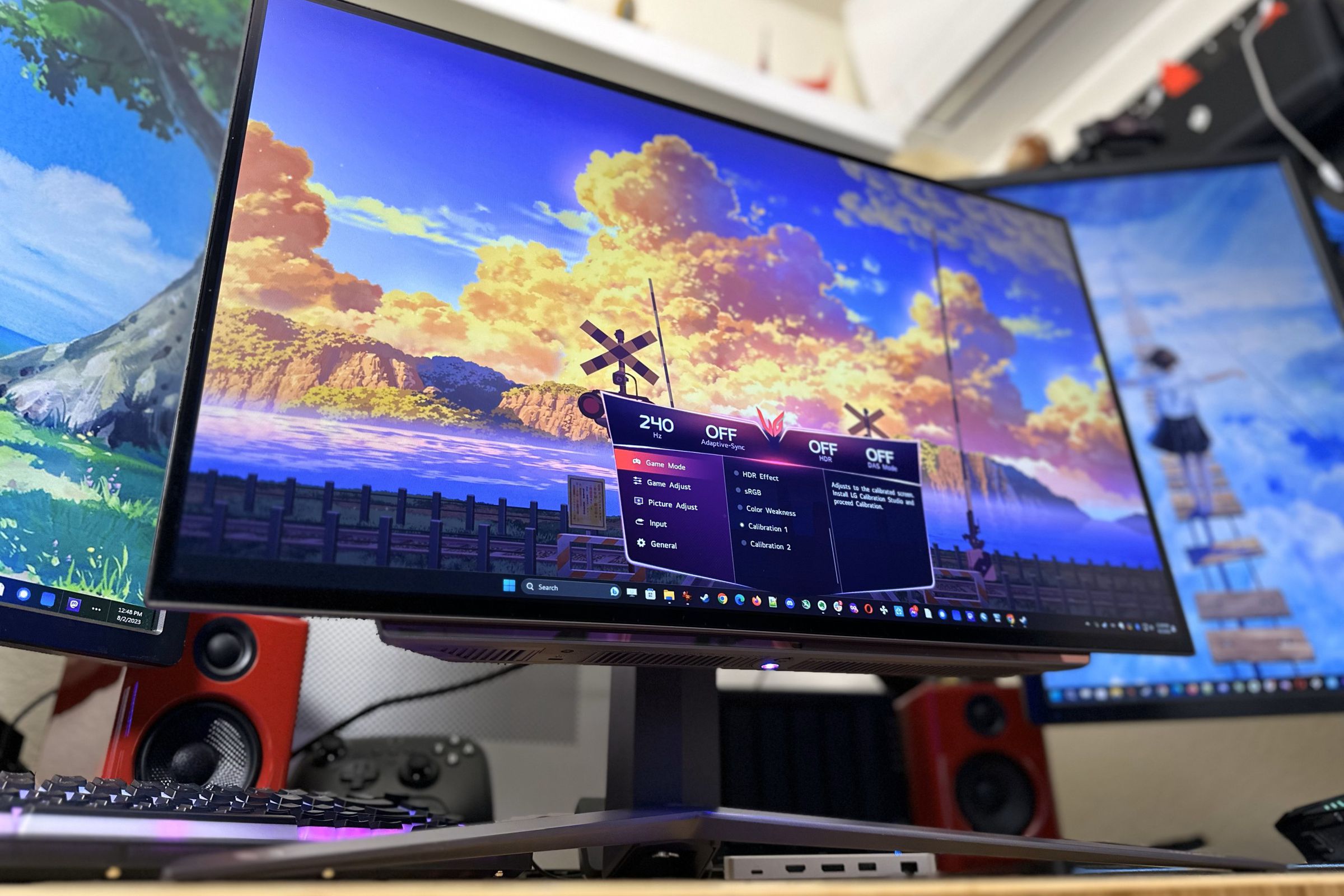 A bright, colorful OLED monitor with a big V-shaped stand underneath, flanked by two other monitors in portrait mode. The main monitor has a colorful sky on screen with big orange sunset-lit clouds and an on-screen display showing it’s running at 240Hz.