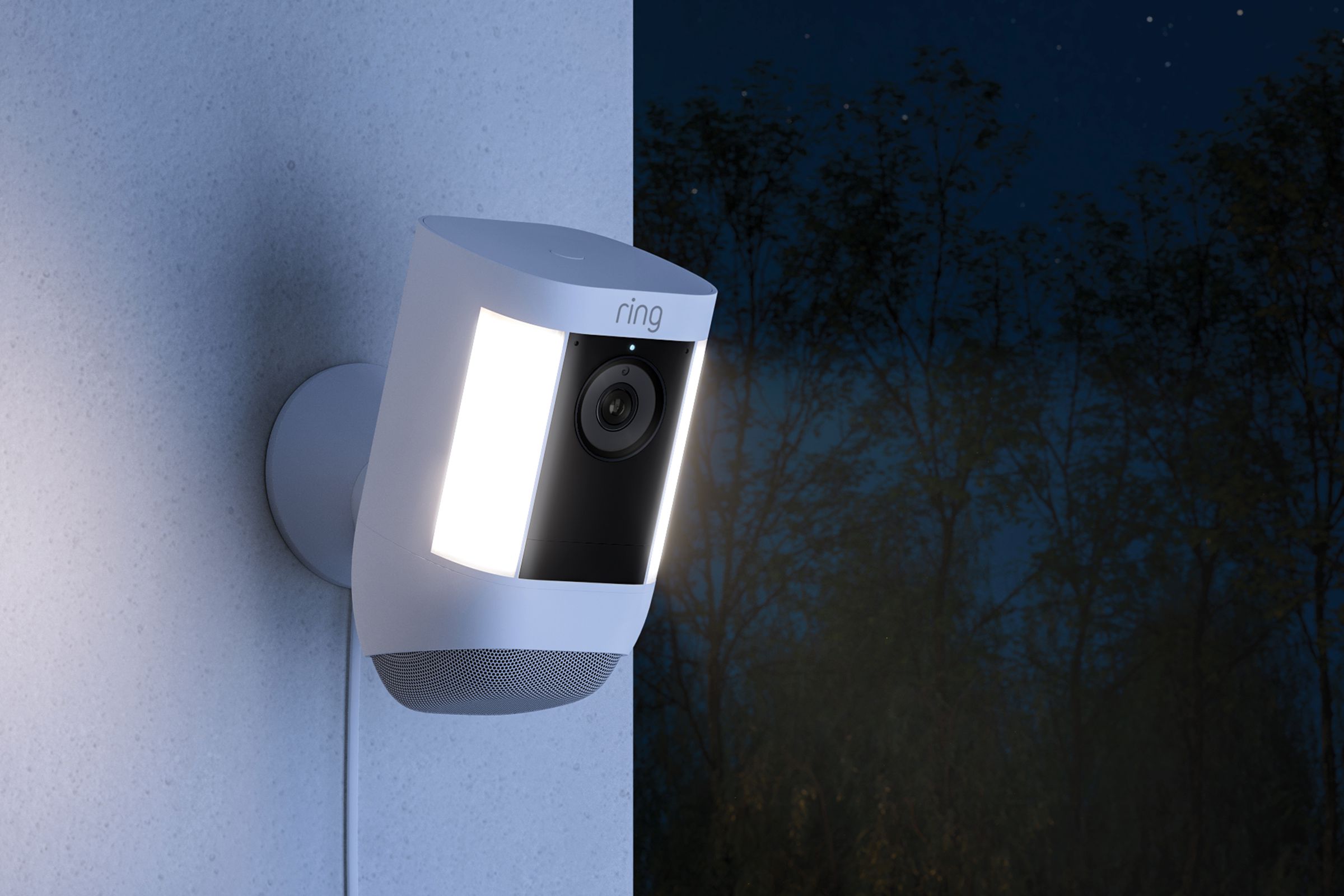 Ring’s spotlight camera is installed on an outdoor white wall with a cable running straight down. Off the wall is a dark wooded background, and the camera has two lights on either side of its body on.