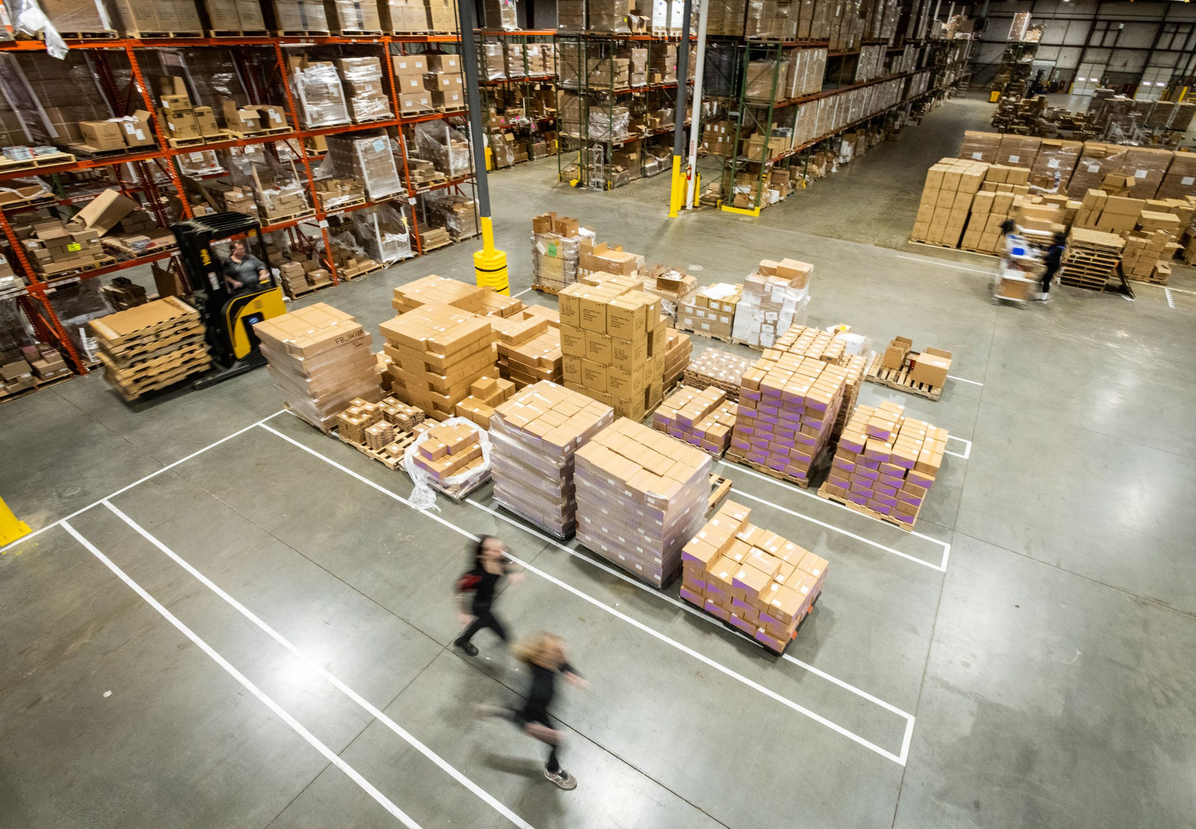Bird’s eye view of a large warehouse floor with boxes stacked on the floor and shelves are at the edge of the frame filled with boxes.