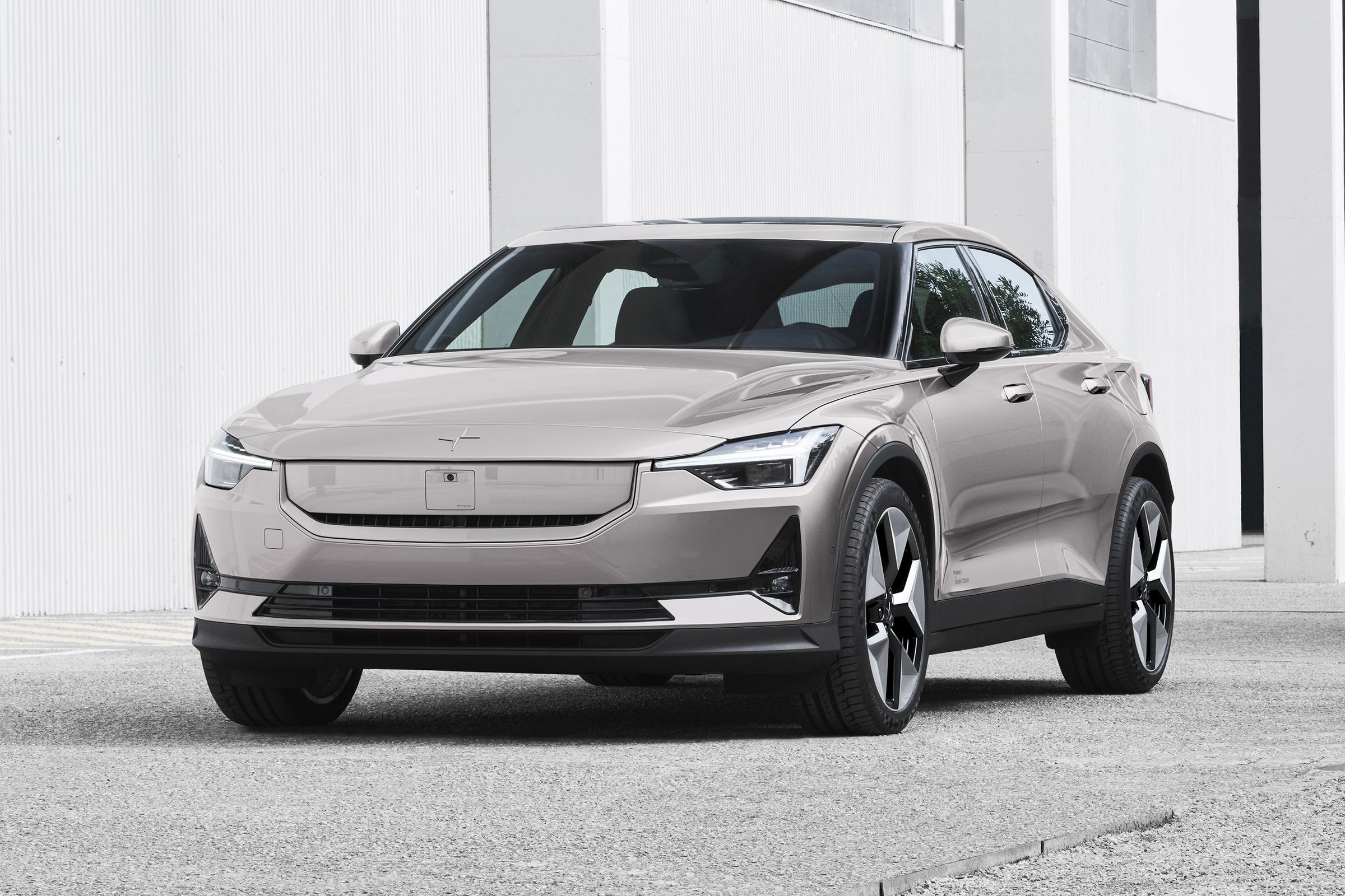 A silver- and beige-colored Polestar 2 is parked on concrete with a white building in the back. The front and driver’s side of the car are visible with a new front that has a body-colored panel covering the grille.