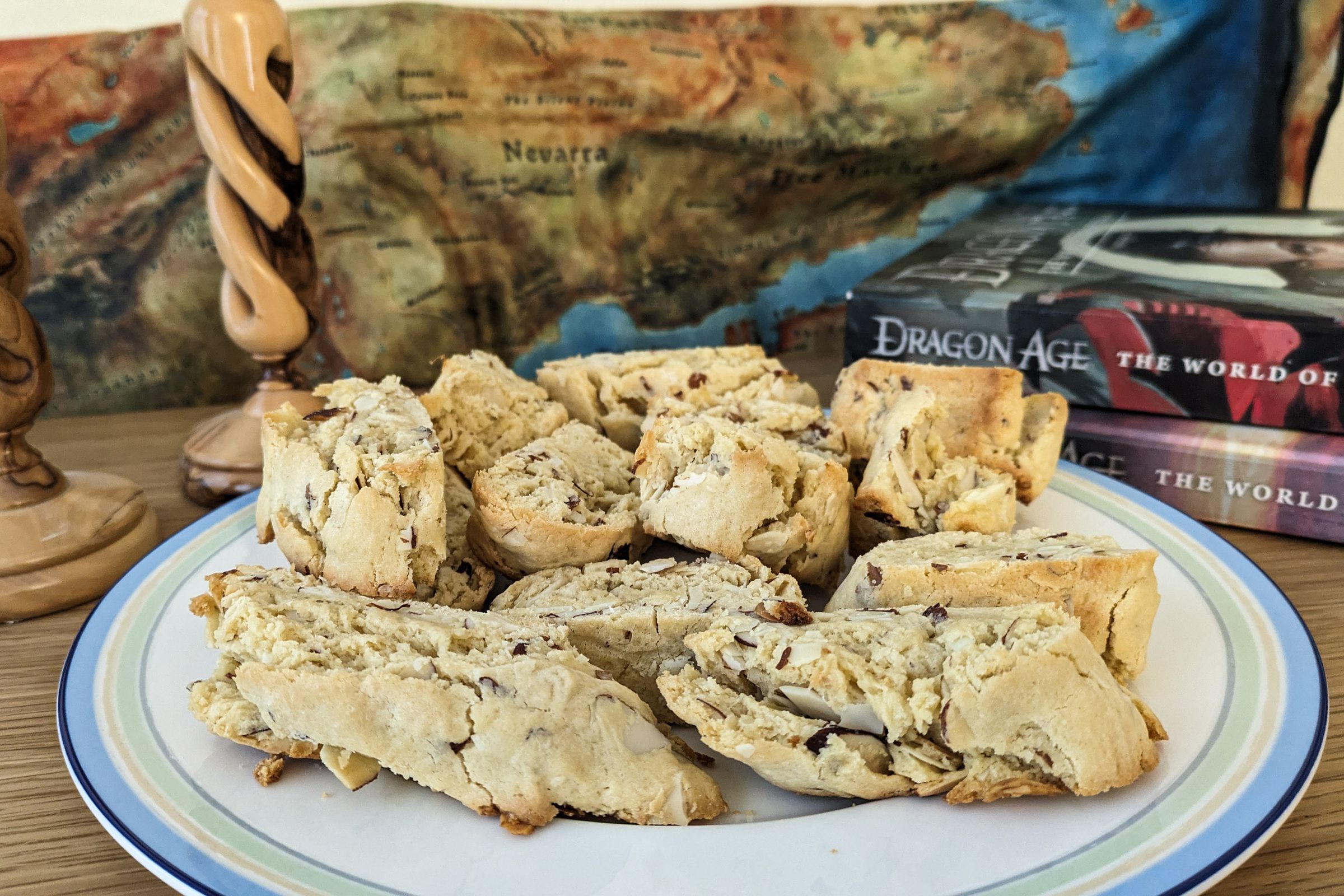 A pile of biscotti-like pastries sitting on a plate in front of a map of Thedas and a pair of wooden candlesticks.