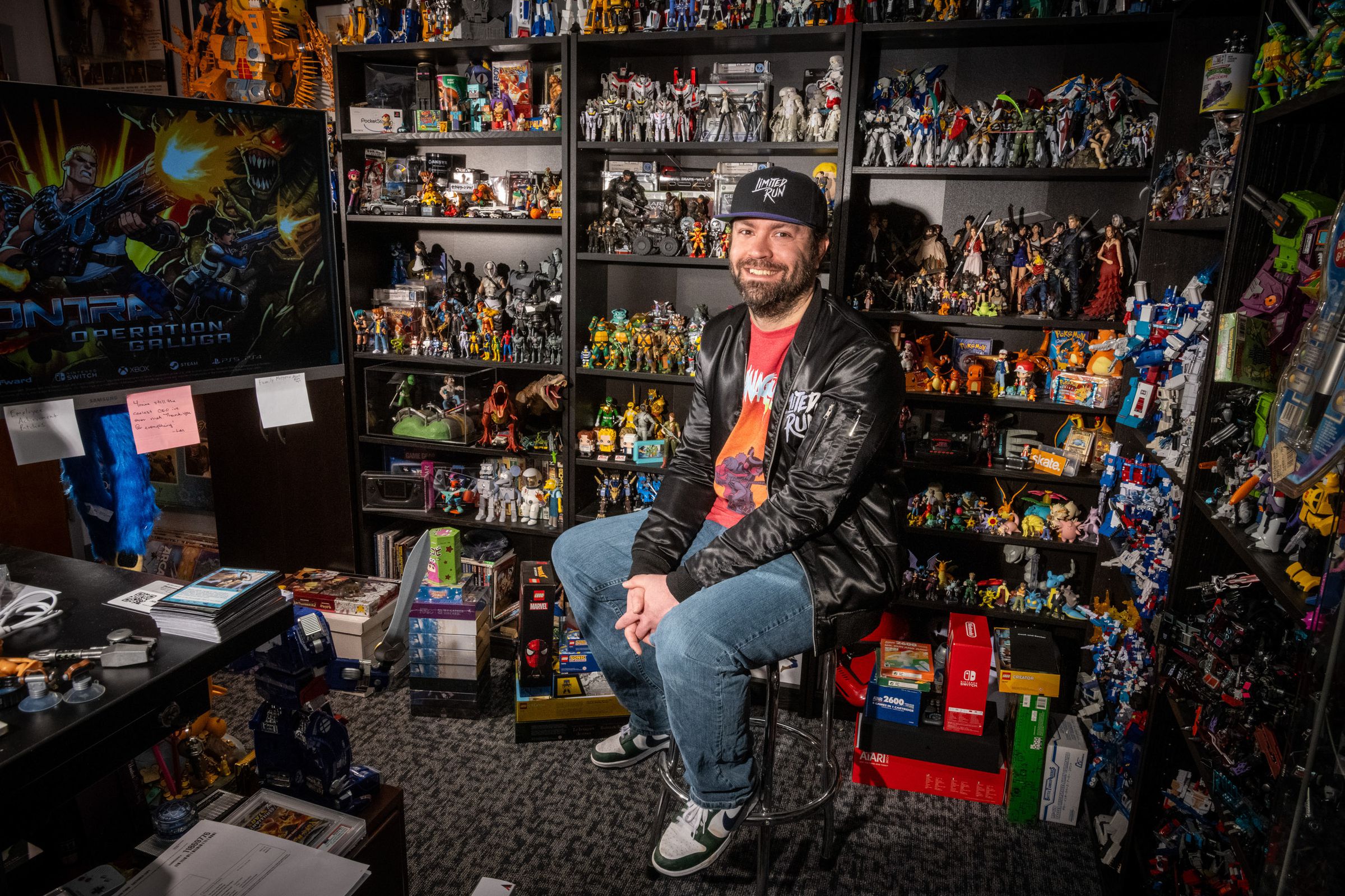 A man sits on a stool in the corner of a room of shelving units that are filled with different collectable figurines. 