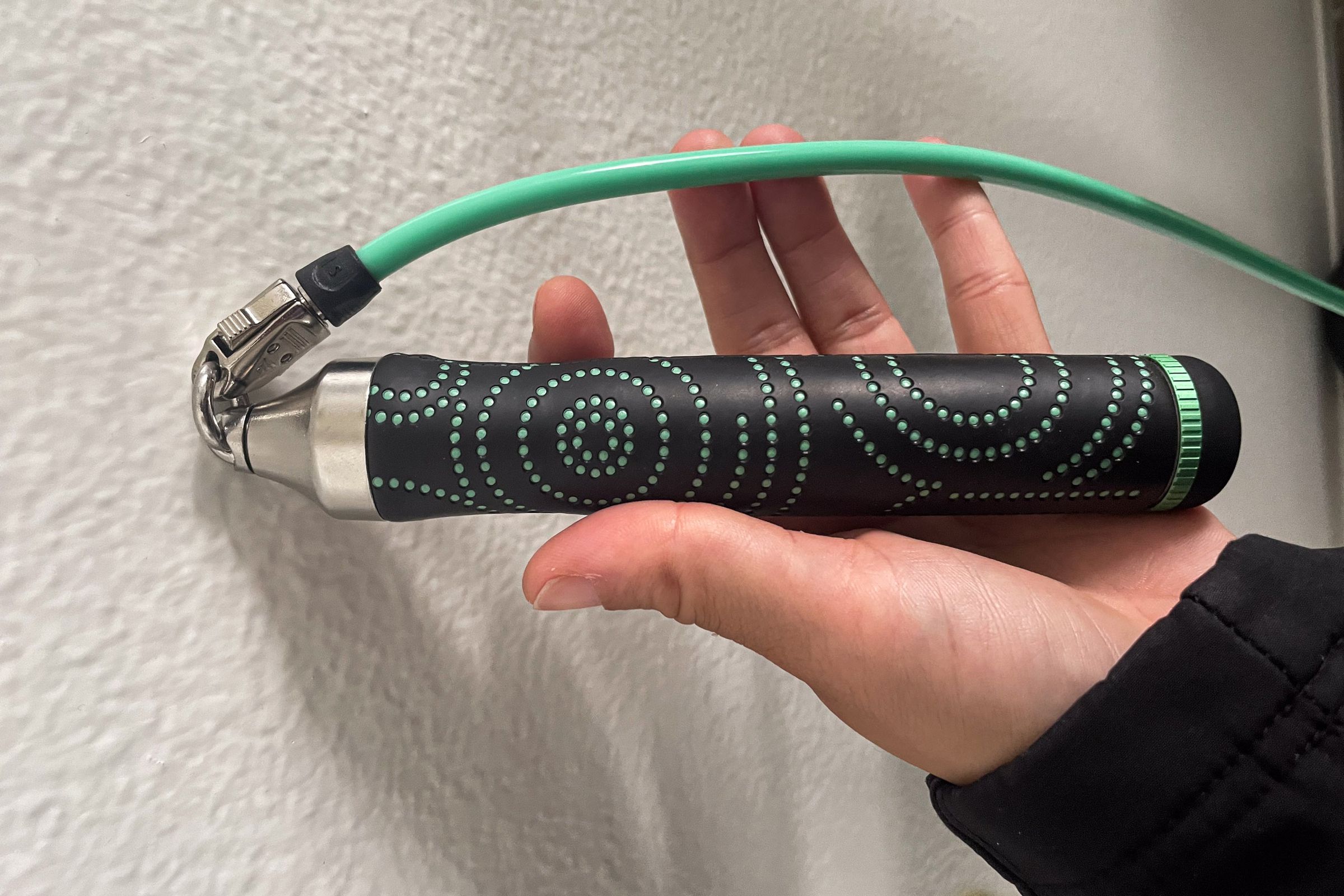 A hand holding a set of black jump-rope handles with green squiggly lines, steel interconnects, and a green rope connecting them.
