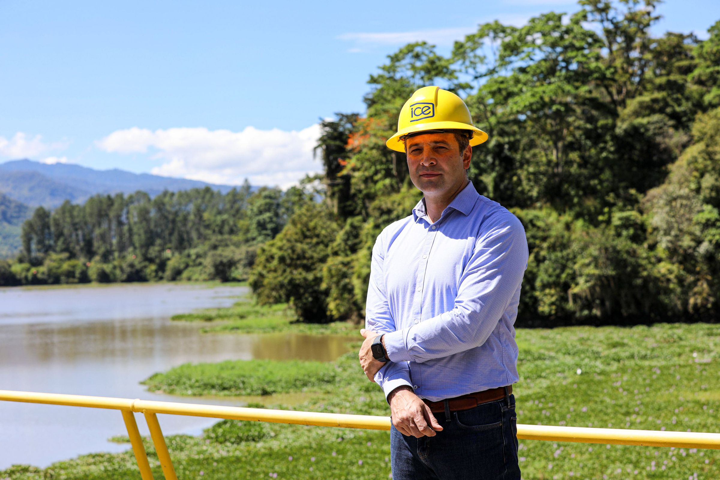 A man wearing a hard hat stands with a hydroelectric reservoir behind him.