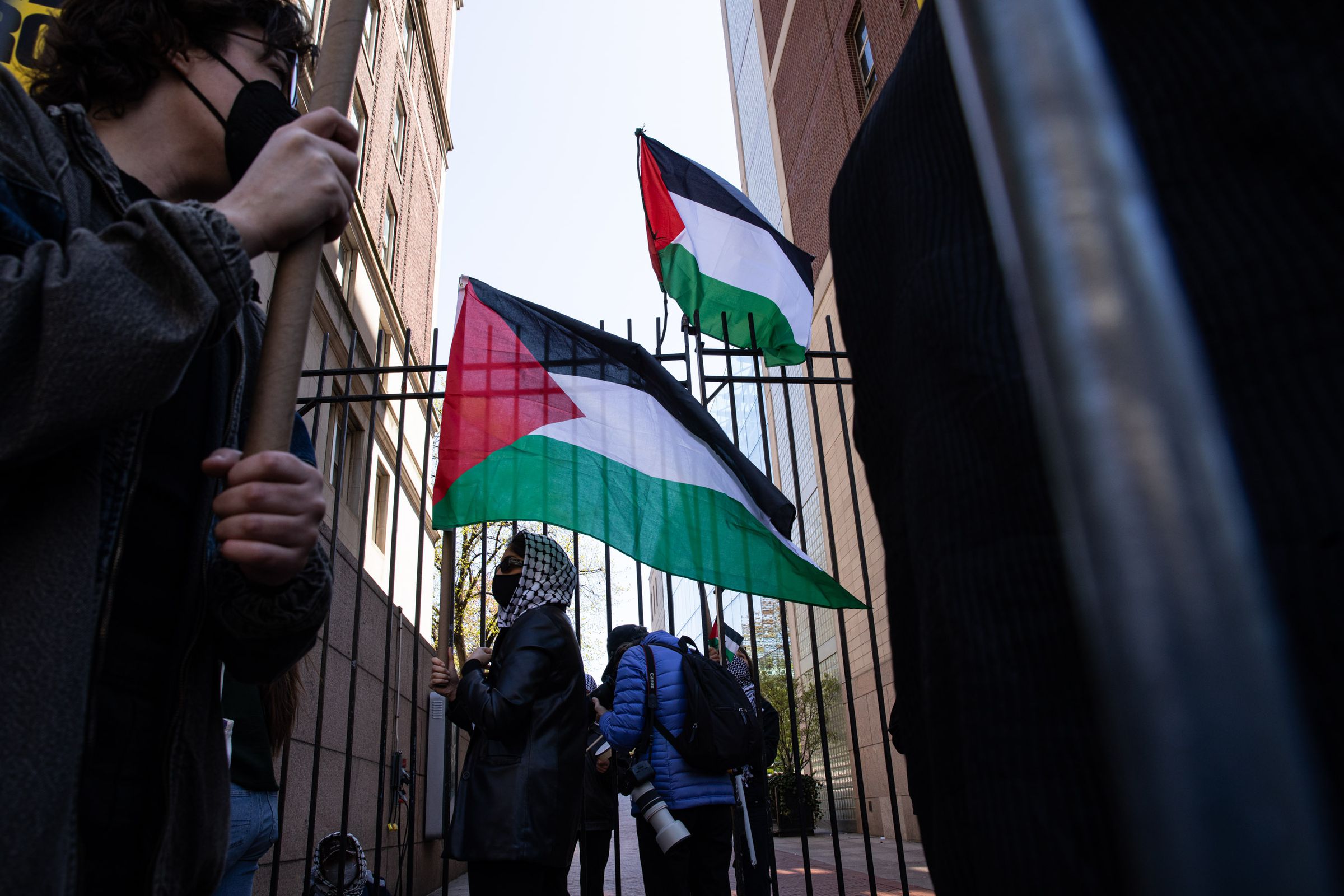 A student wears a mask and kaffiyeh and carries a Palestinian flag.