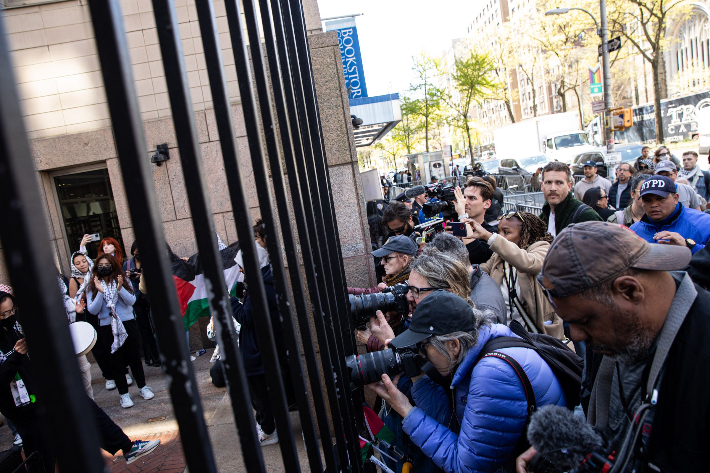 A crowd of photographers presses in at the wrought-iron gates of the university. Behind the fence, students demonstrate wearing kaffiyehs and waving Palestinian flags.