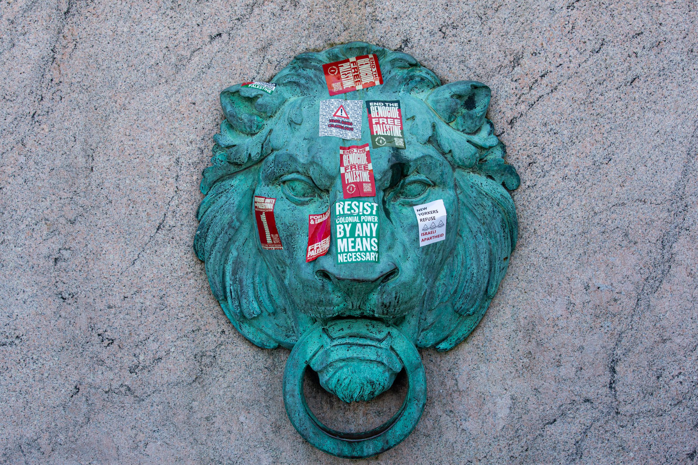 A photo of a patinaed bronze lion head affixed to marble. The lion head is covered in political stickers, including slogans like “End the Genocide, Free Palestine.”