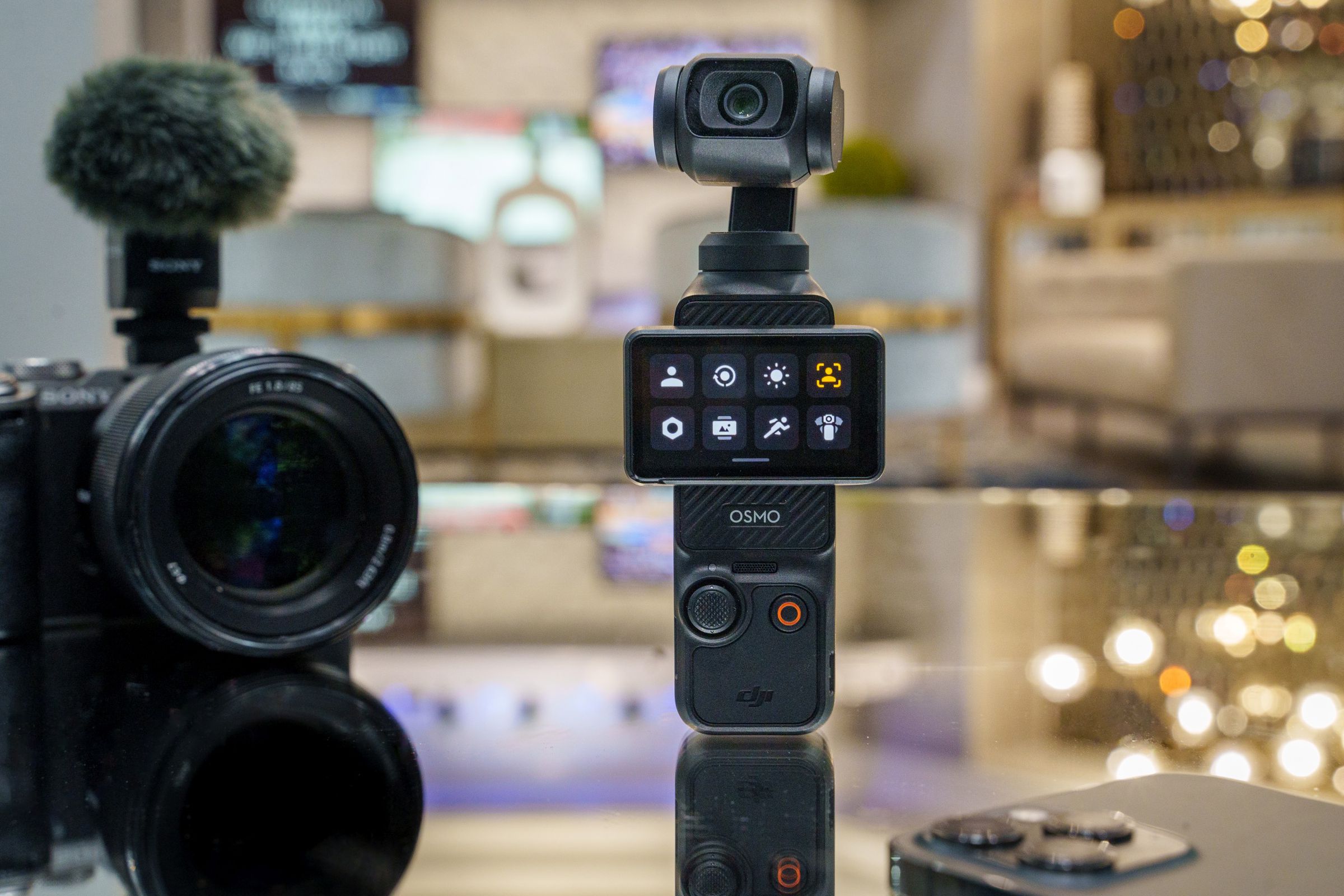 Osmo Pocket 3 mirrored on a glass table, with an iPhone in the foreground and a Sony mirrorless camera in the background