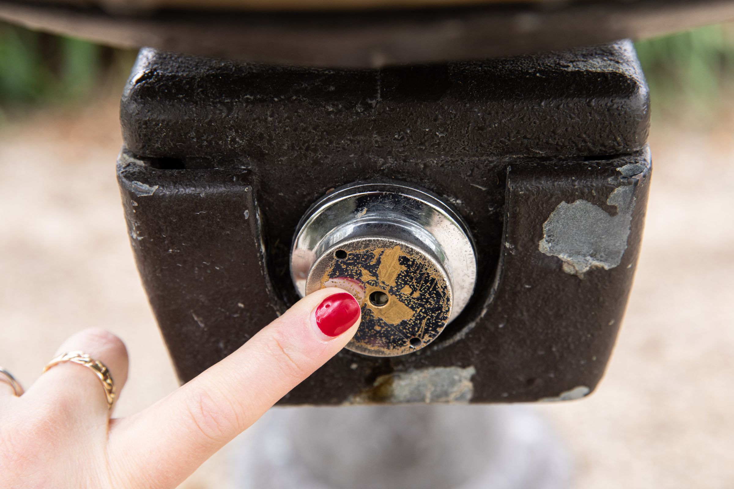 A finger with red nail polish presses a worn metal disc of a drinking fountain button with three holes