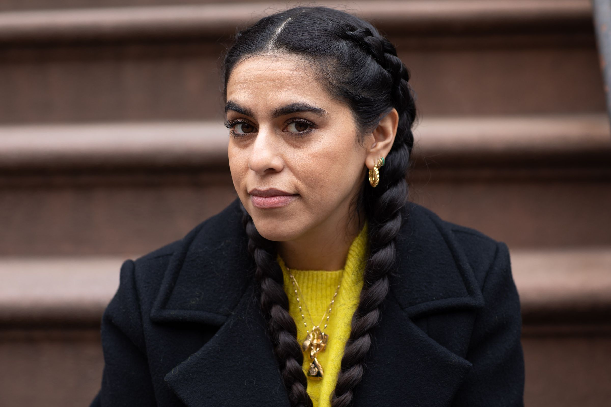 Photo of Mona Chalabi. Her hair is in two braids and she is wearing a black coat and yellow sweater. She is seated on the front steps of her apartment.