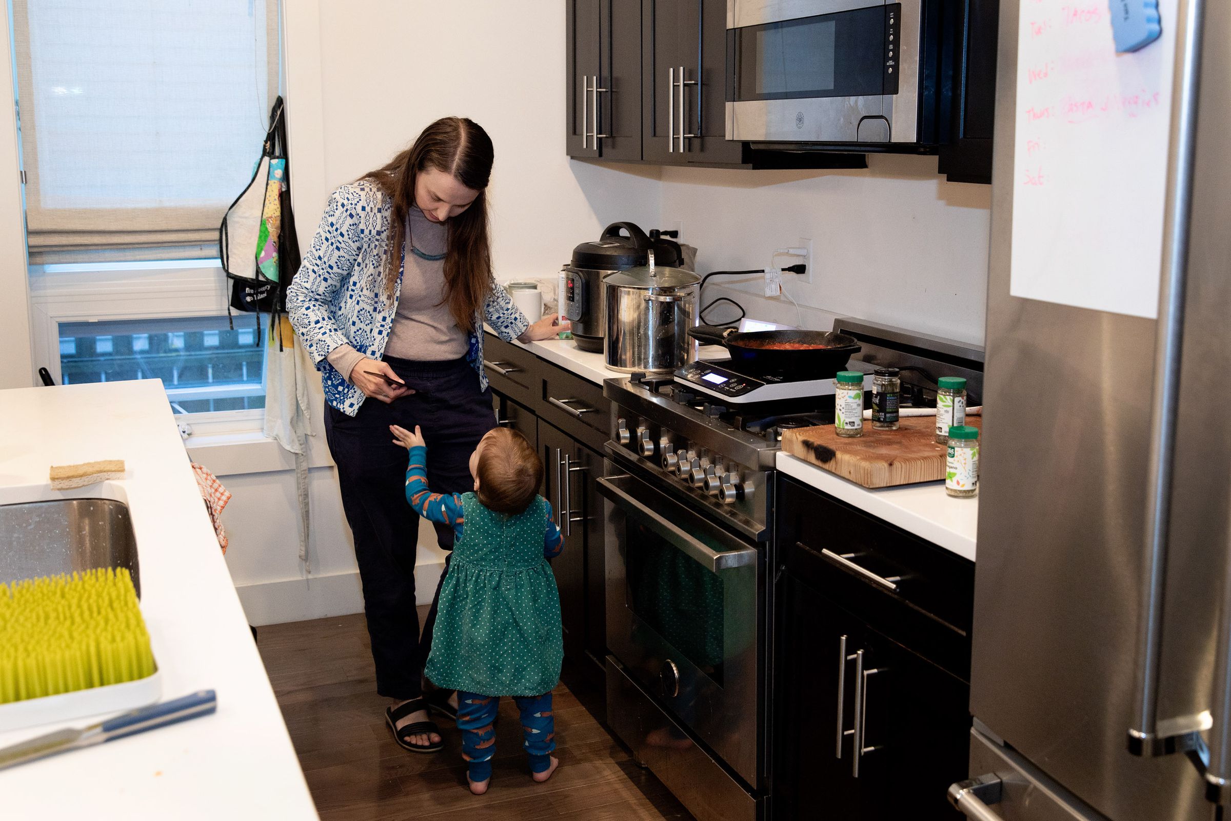 A person with long hair leans over to speak to a toddler who we see from behind. They are standing in a narrow kitchen in front of the stove.