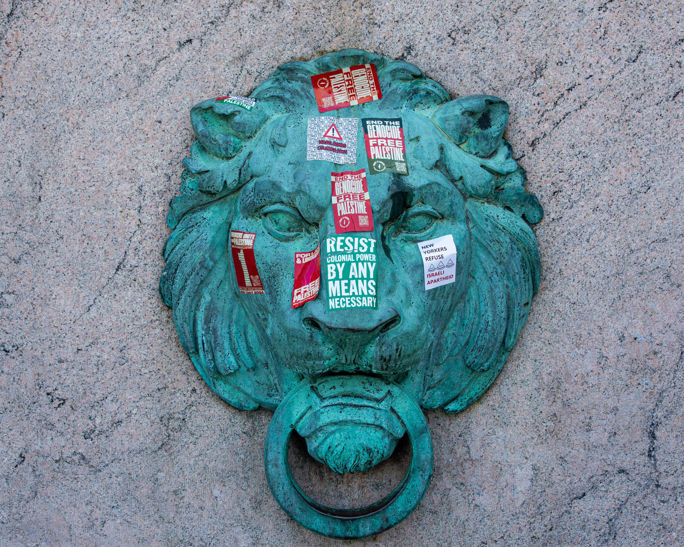 A photo of a patinaed bronze lion head affixed to marble. The lion head is covered in political stickers, including slogans like “End the Genocide, Free Palestine.”