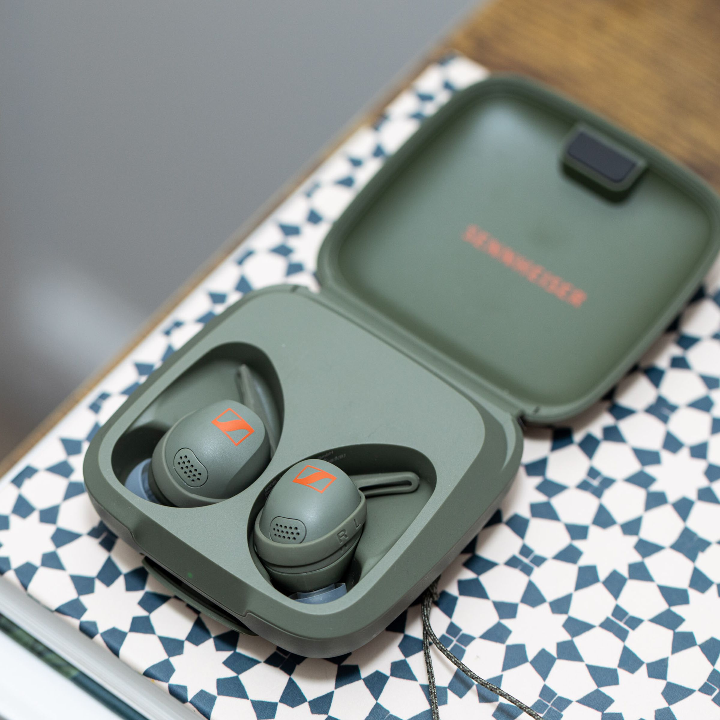 Olive-colored Sennheiser Momentum Sport buds in their case on top of a patterned book.
