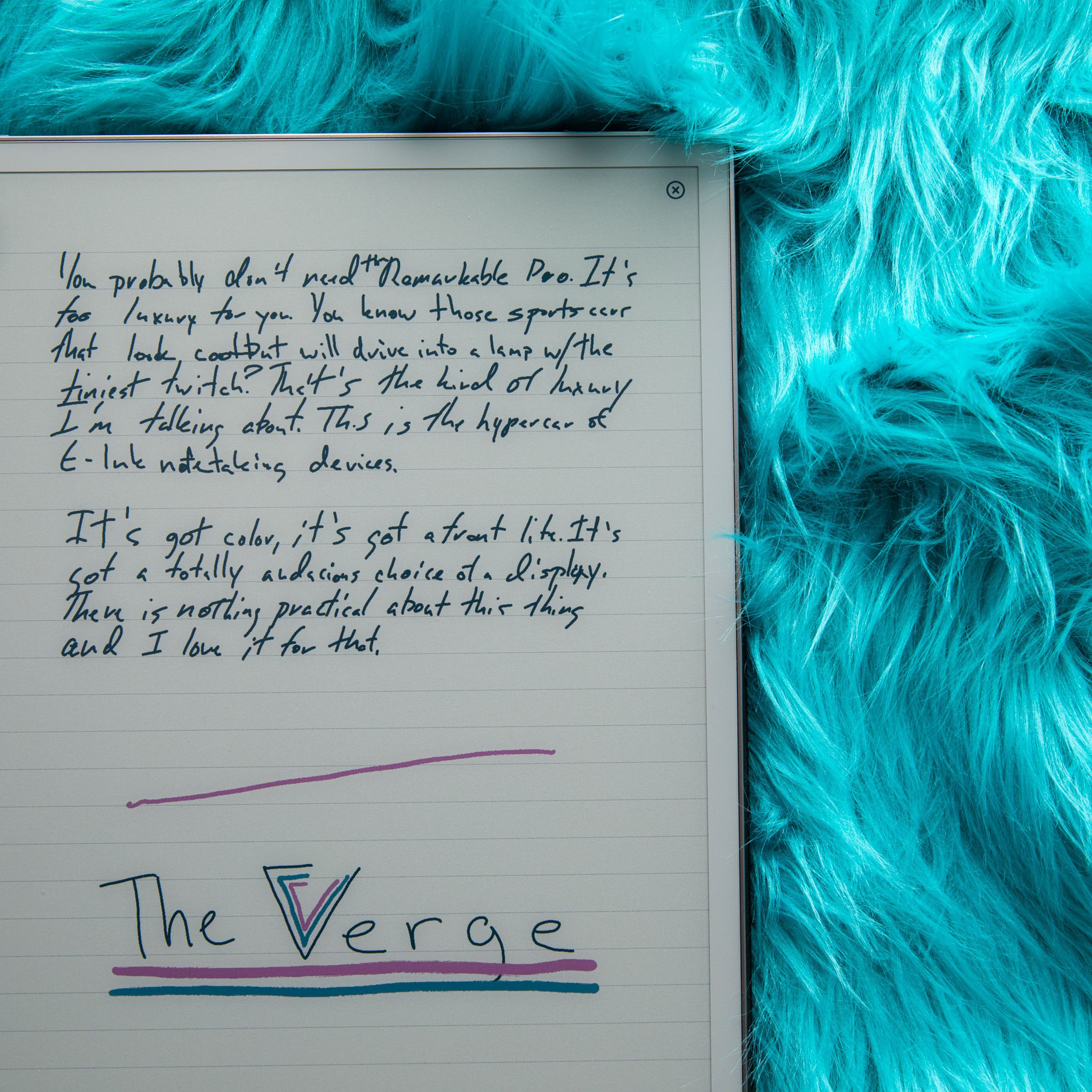 A picture of a color E Ink display resting on what might be the pelt of an electric-blue Wookiee. Handwritten notes in black “ink” cover the top half of the display, and a The Verge logo is drawn on the lower half, with a magenta line above it, the V in “Verge” done in three concentric V’s of black, pink, and blue, and “The Verge” underlined once in blue and once in magenta.