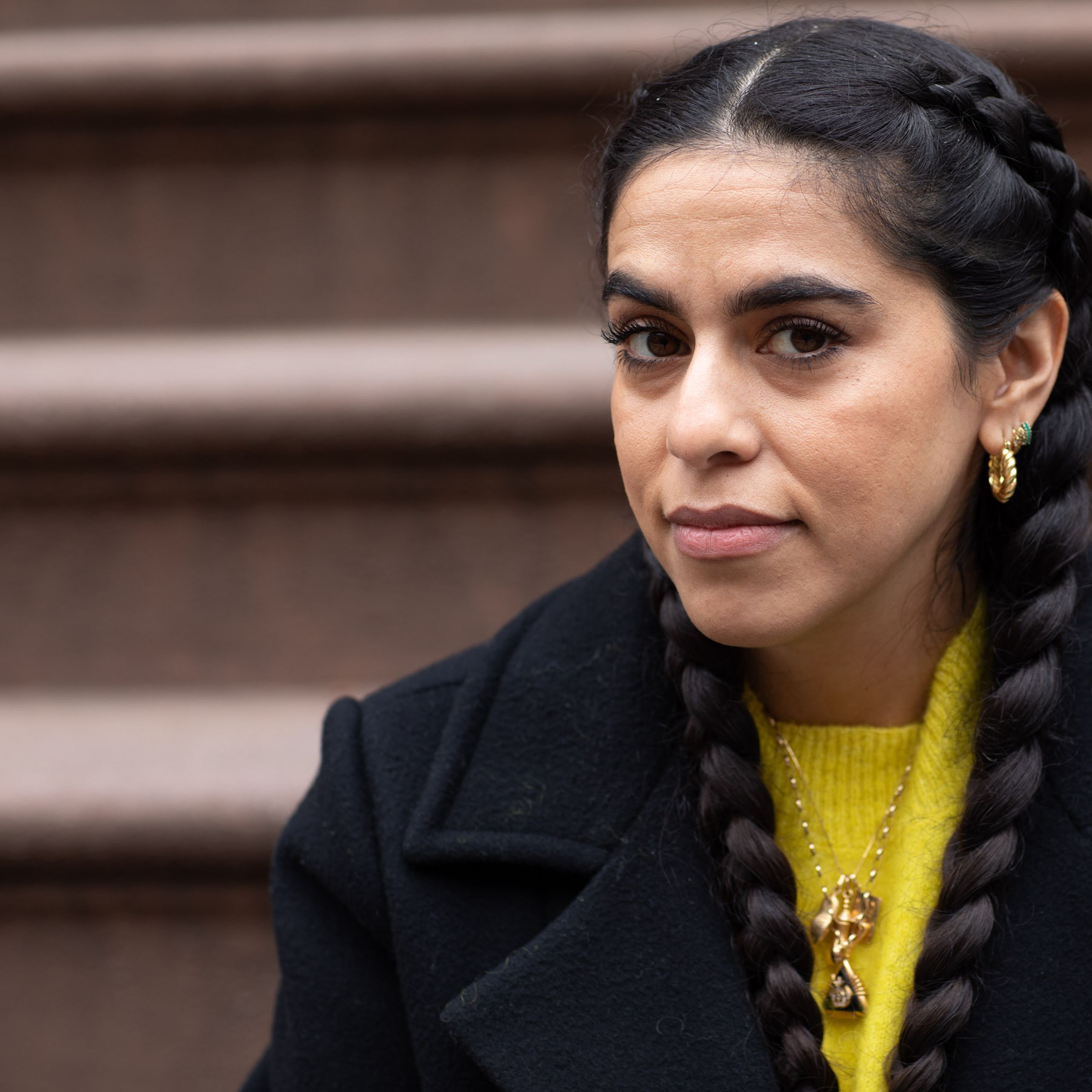 Photo of Mona Chalabi. Her hair is in two braids and she is wearing a black coat and yellow sweater. She is seated on the front steps of her apartment.