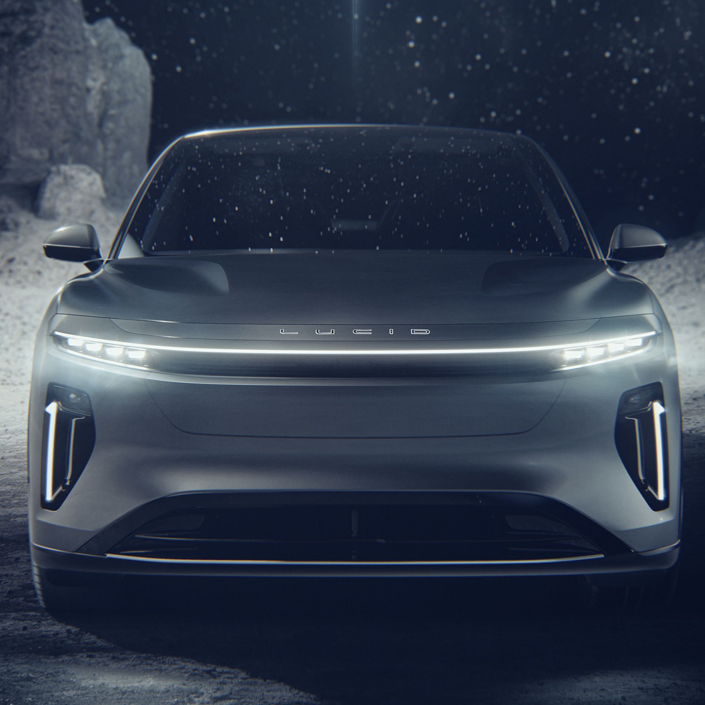 Front end of the Lucid Gravity — it’s an SUV with an LED bar light and LEF headlights with a tall nose that has no grille, a large air intake in the bottom, and air holes on both sides of the bumper. The car is in a rocky and dark environment.