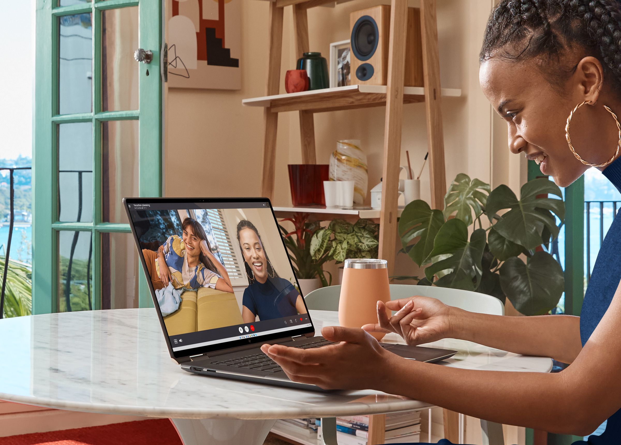 A user participates in a video call on the HP Spectre x360 16. The laptop is seen from the side and sits on a marble table. A hydroflask is to its right, as is a shelf populated with two potted plants, a stack of books, a number of glass cups and jars, a pencil pot, and a speaker.