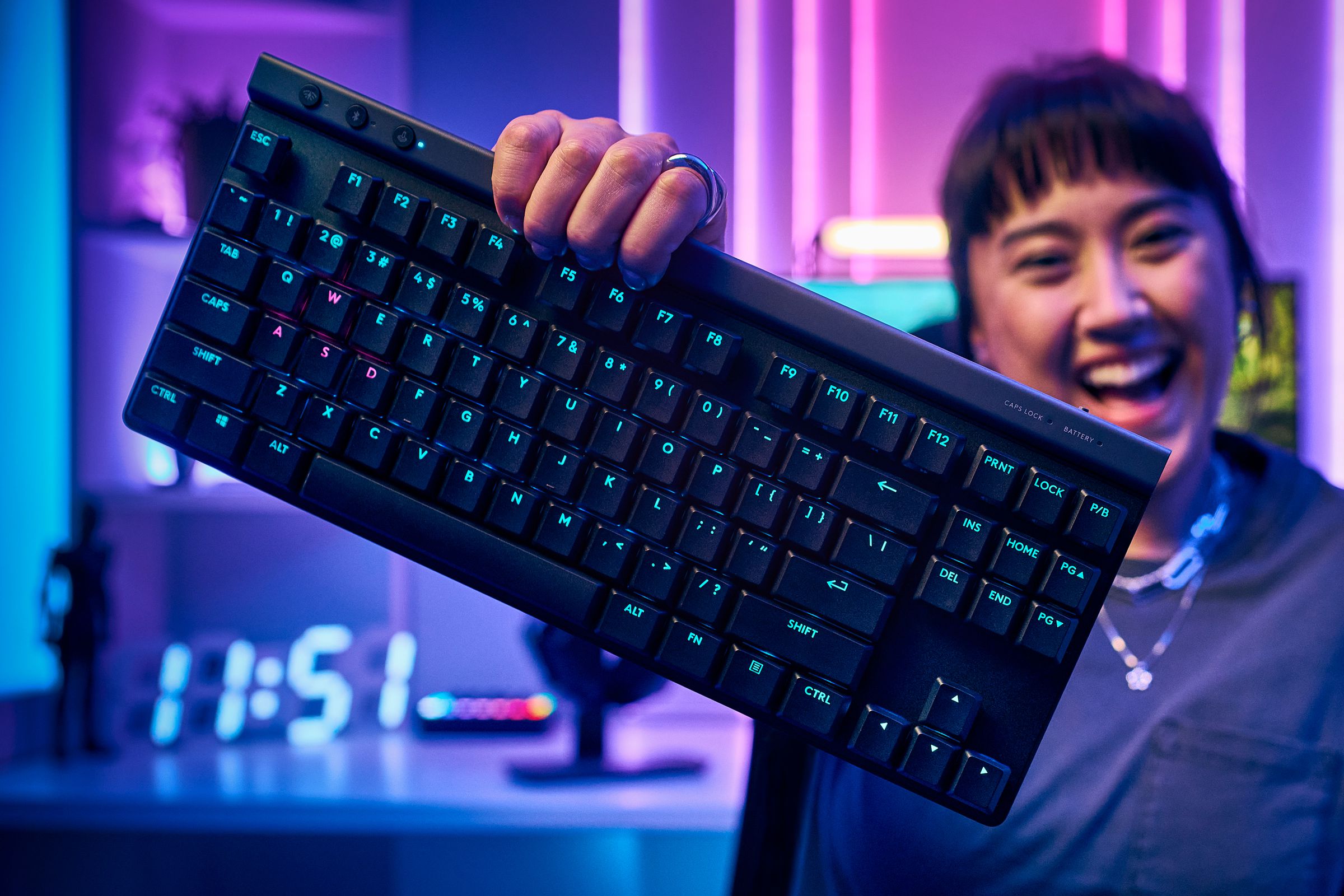 A user holds the Logitech G515 keyboard in one hand in front a computer setup featuring neon lighting.