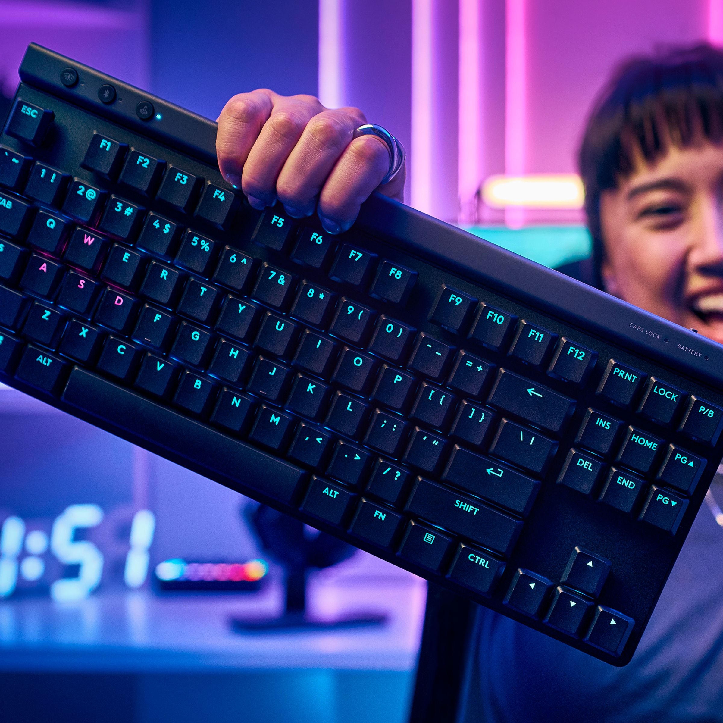 A user holds the Logitech G515 keyboard in one hand in front a computer setup featuring neon lighting.