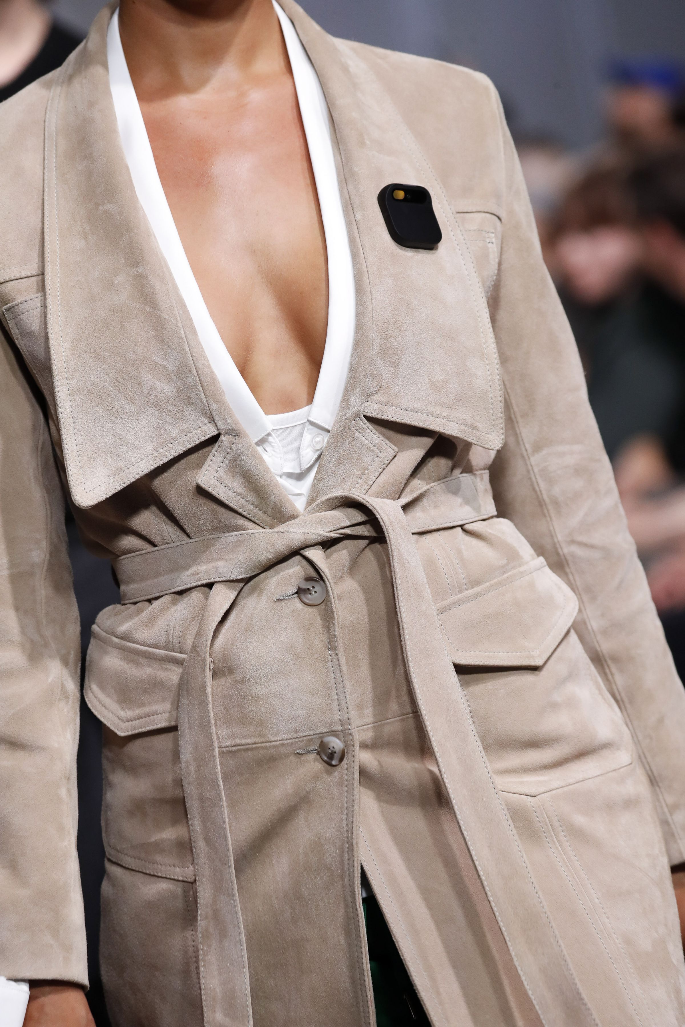 A close-up of a model, from the neck down, showing the Humane AI pin on their tan jacket’s broad lapel.