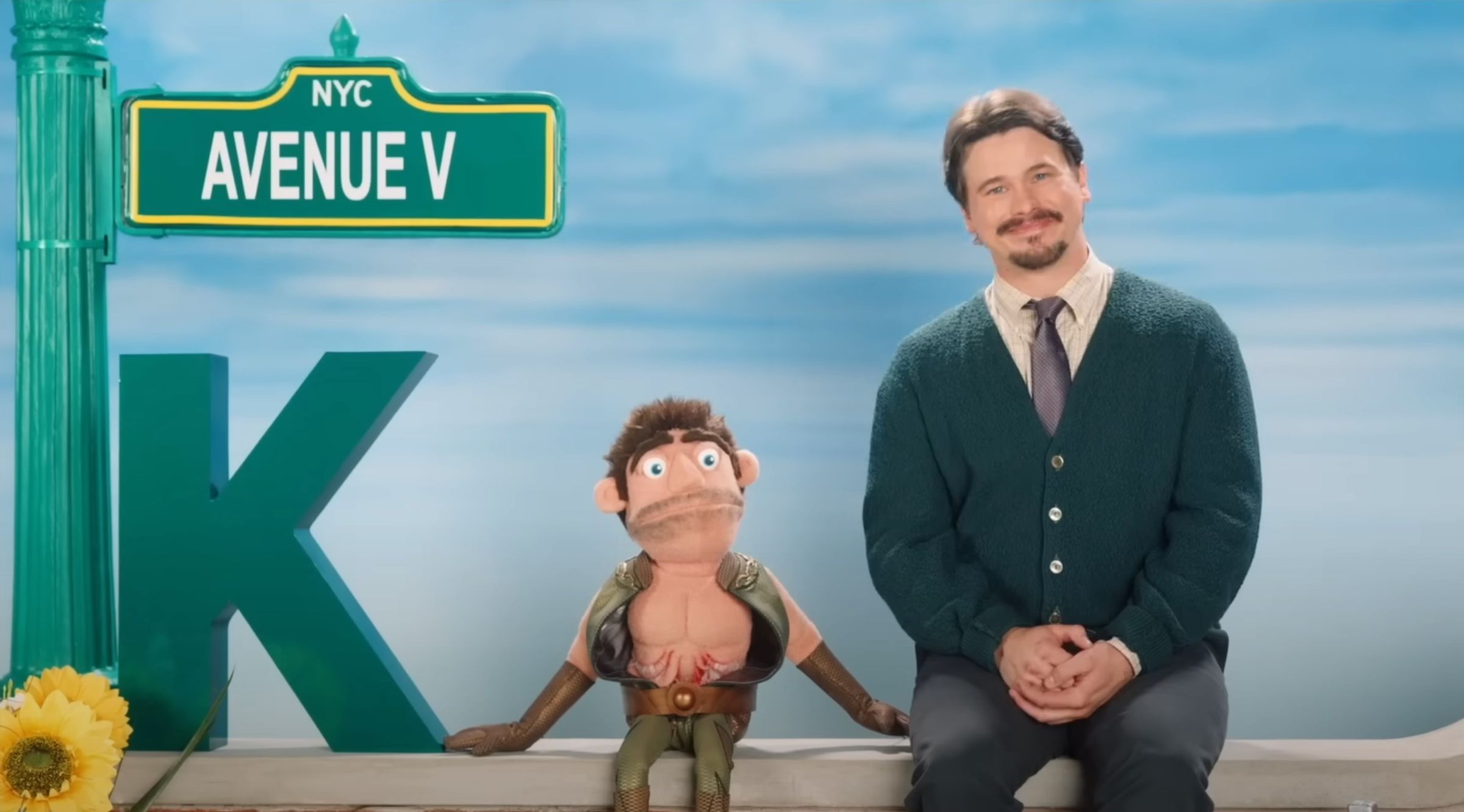 A screenshot taken from the Gen V trailer. A man in a green cardigan is sitting next to a puppet of The Deep from The Boys.