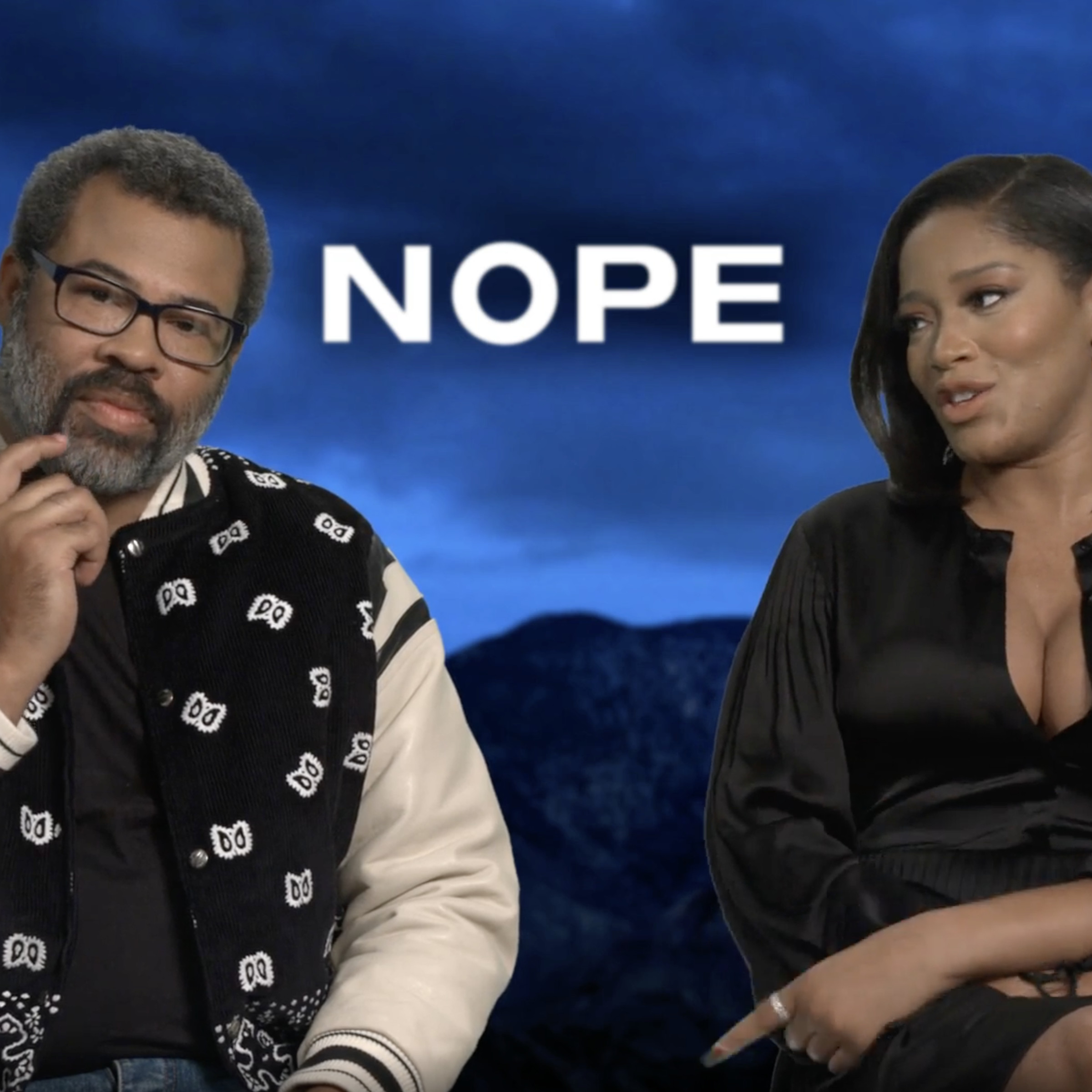 Jordan Peele wearing a black bomber jacket with white sleeves and a paisley-like pattern and Keke Palmer in a silk dress. Both are sitting in front of a screenshot from the movie Nope depicting an overcast sky and mountains.