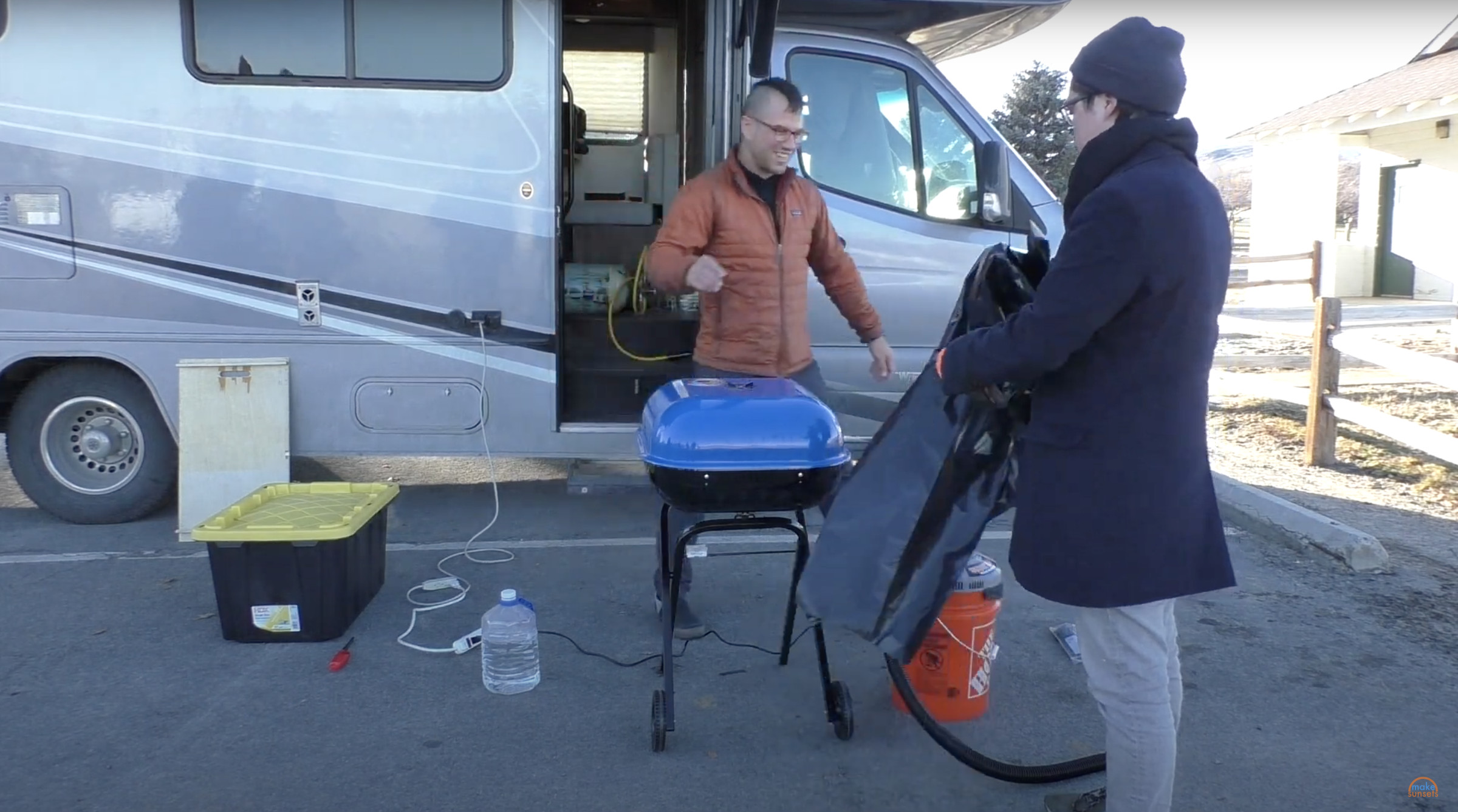 Two guys stand in a parking lot with a small blue charcoal grill between them.