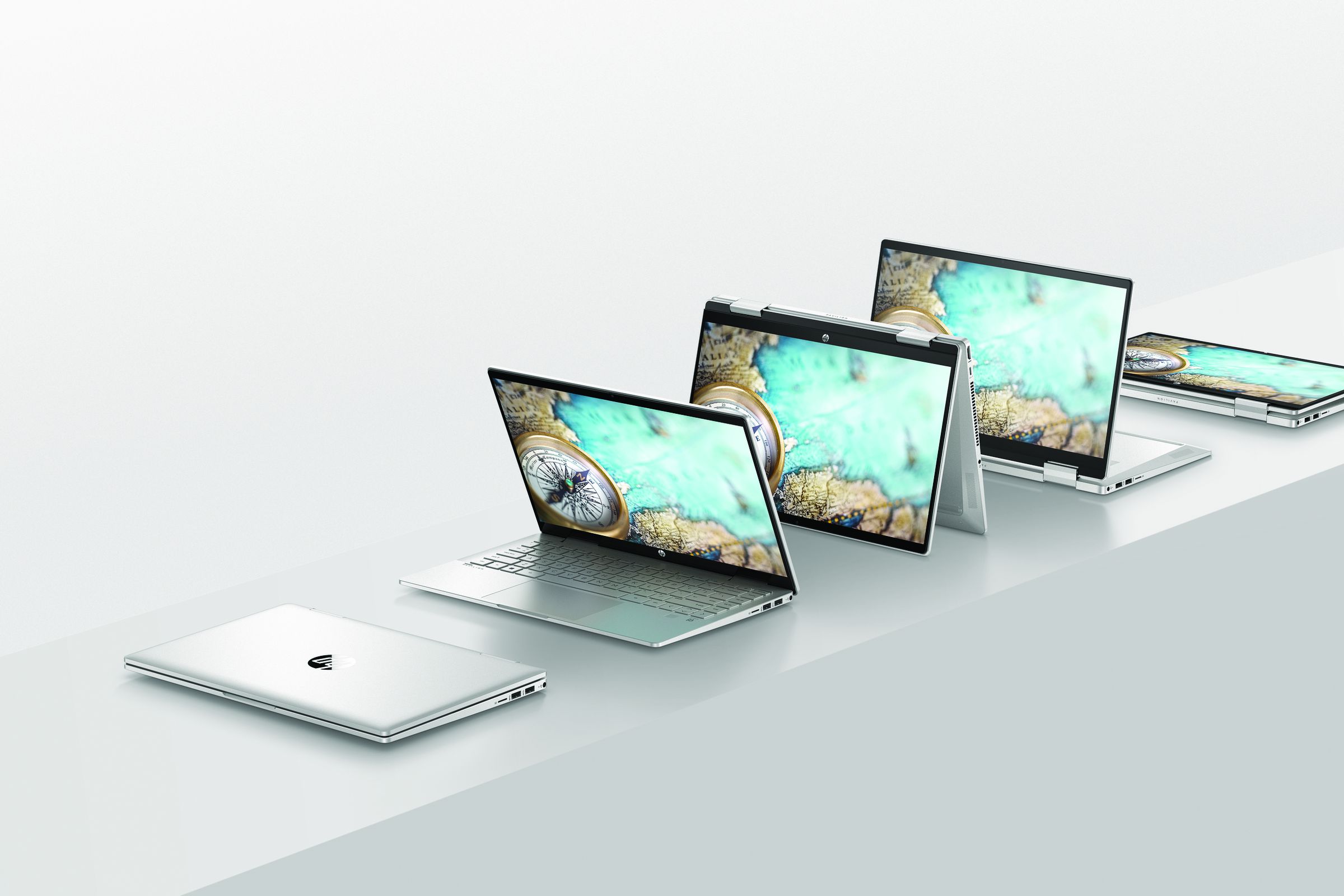 Five HP Pavilion x360 14 models lined up on a white counter in cloesd, clamshell, tent, backwards, and tablet modes respectively. The screen displays a bird’s-eye view of cliffs and water.
