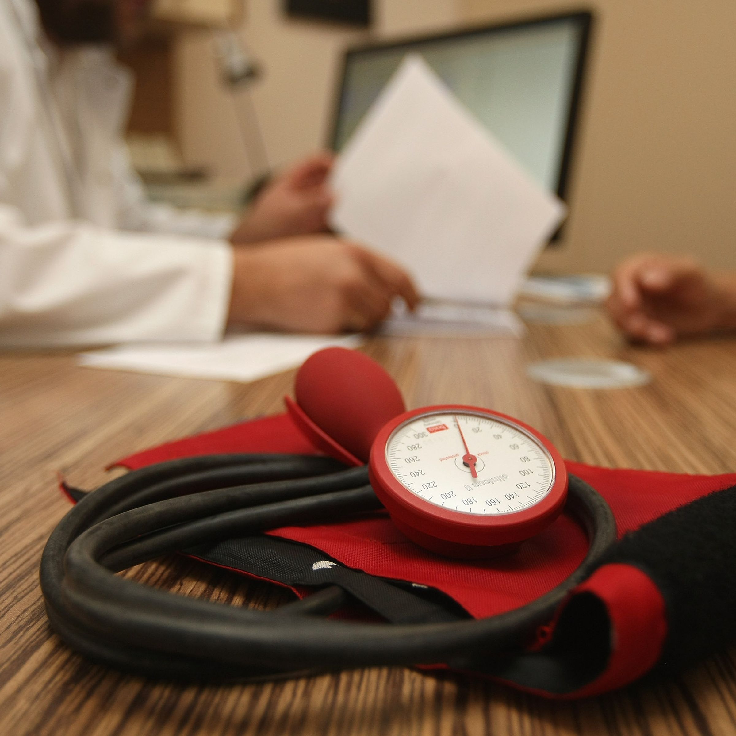 A red blood pressure meter sits on a wooden desk, in the background, a doctor looks over papers. A patient’s arm is also visible on the desk. 