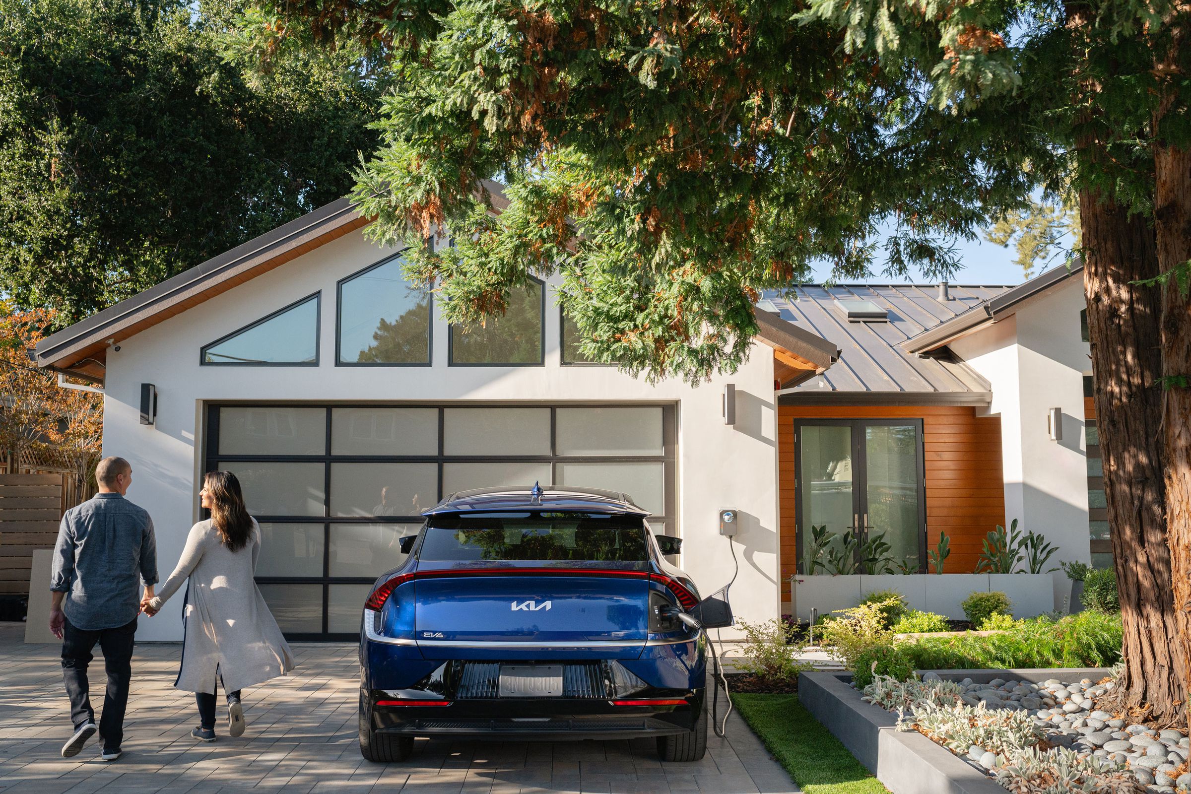 modern house with blue kia ev6 parked in front of garage with charge point charger Hooked up, and two people holding hands walking away from the car