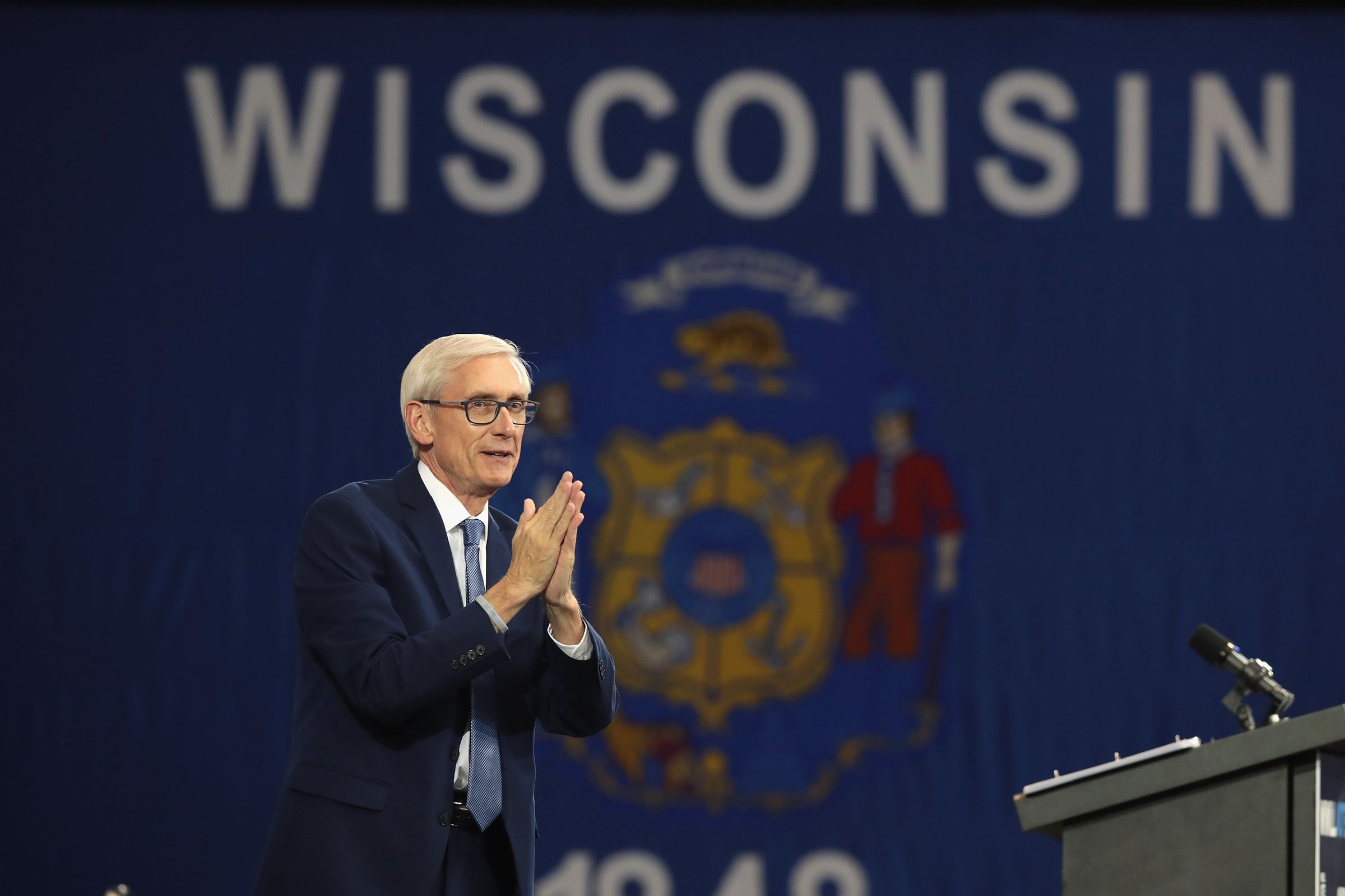 Wisconsin Governor Tony Evers stands in front of an enormous Wisconsin state flag, his palms pressed together