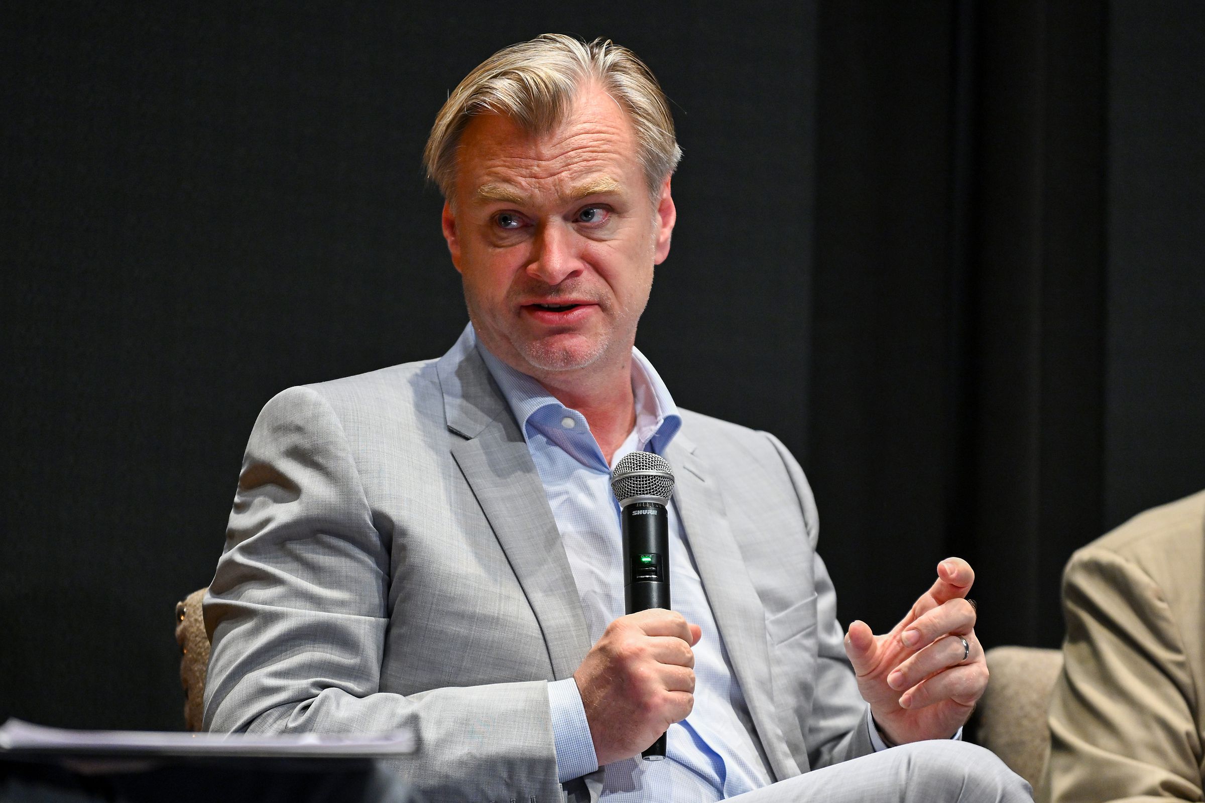 Christopher Nolan, a white man, sits at a panel holding a microphone and speaking.