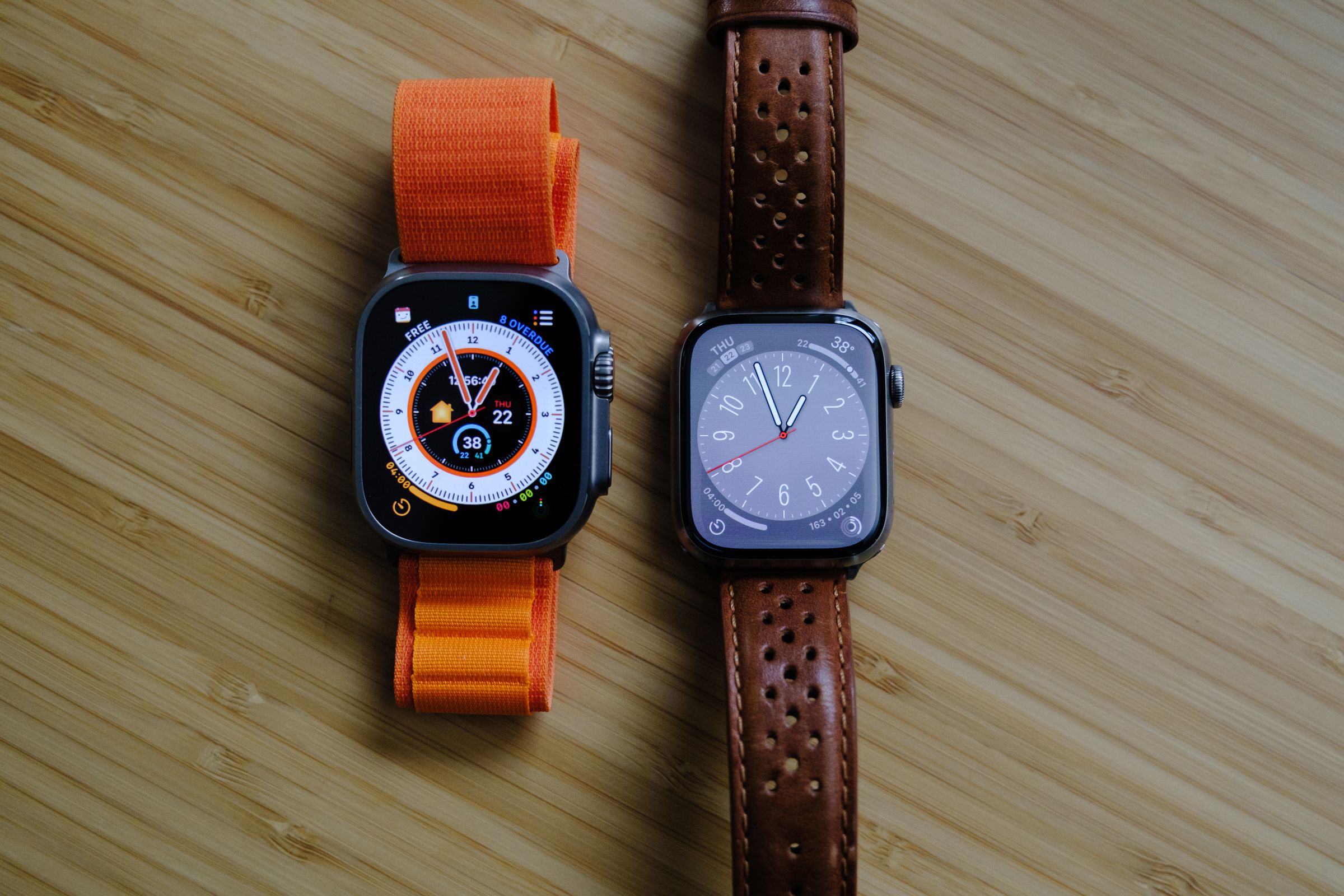 An Apple Watch Ultra next to a Series 7 Edition on a light wood table, viewed from the top.