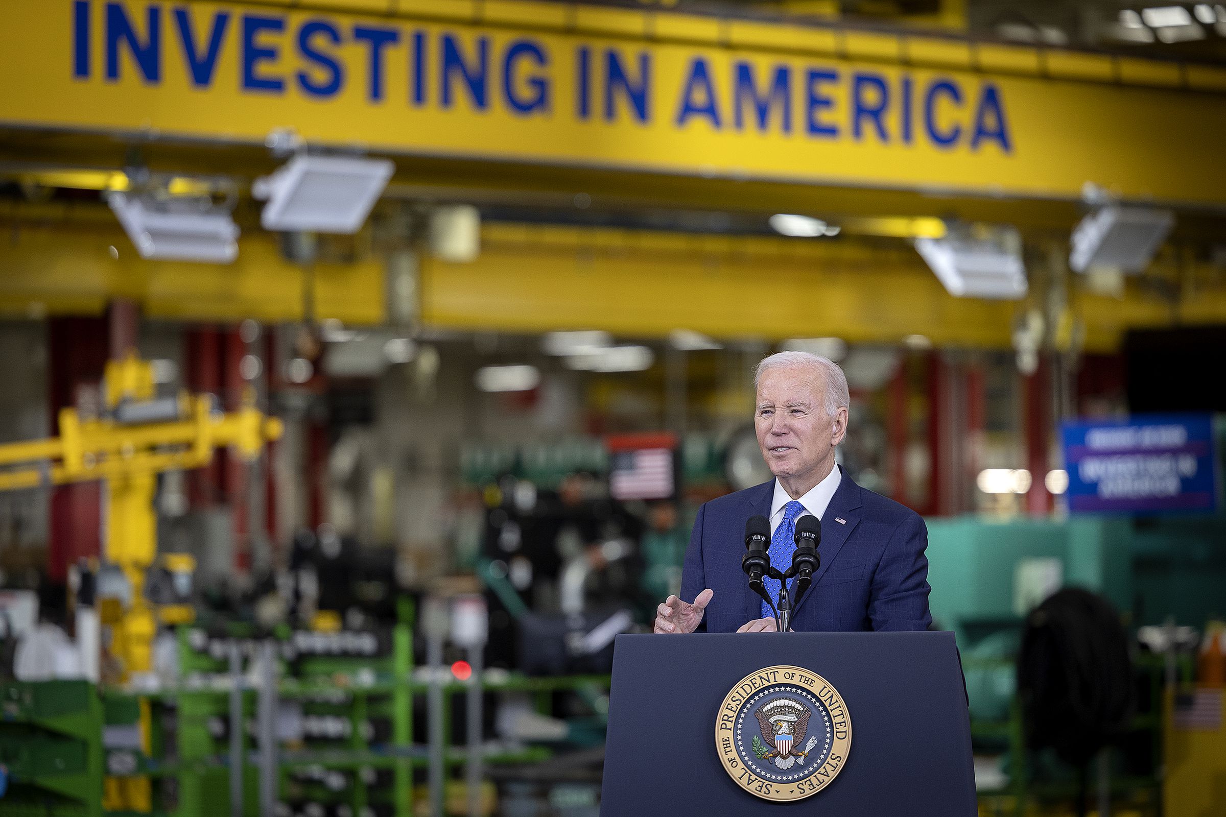 President Joe Biden speaks at a podium in an industrial building with signage that says “investing in America” behind him.