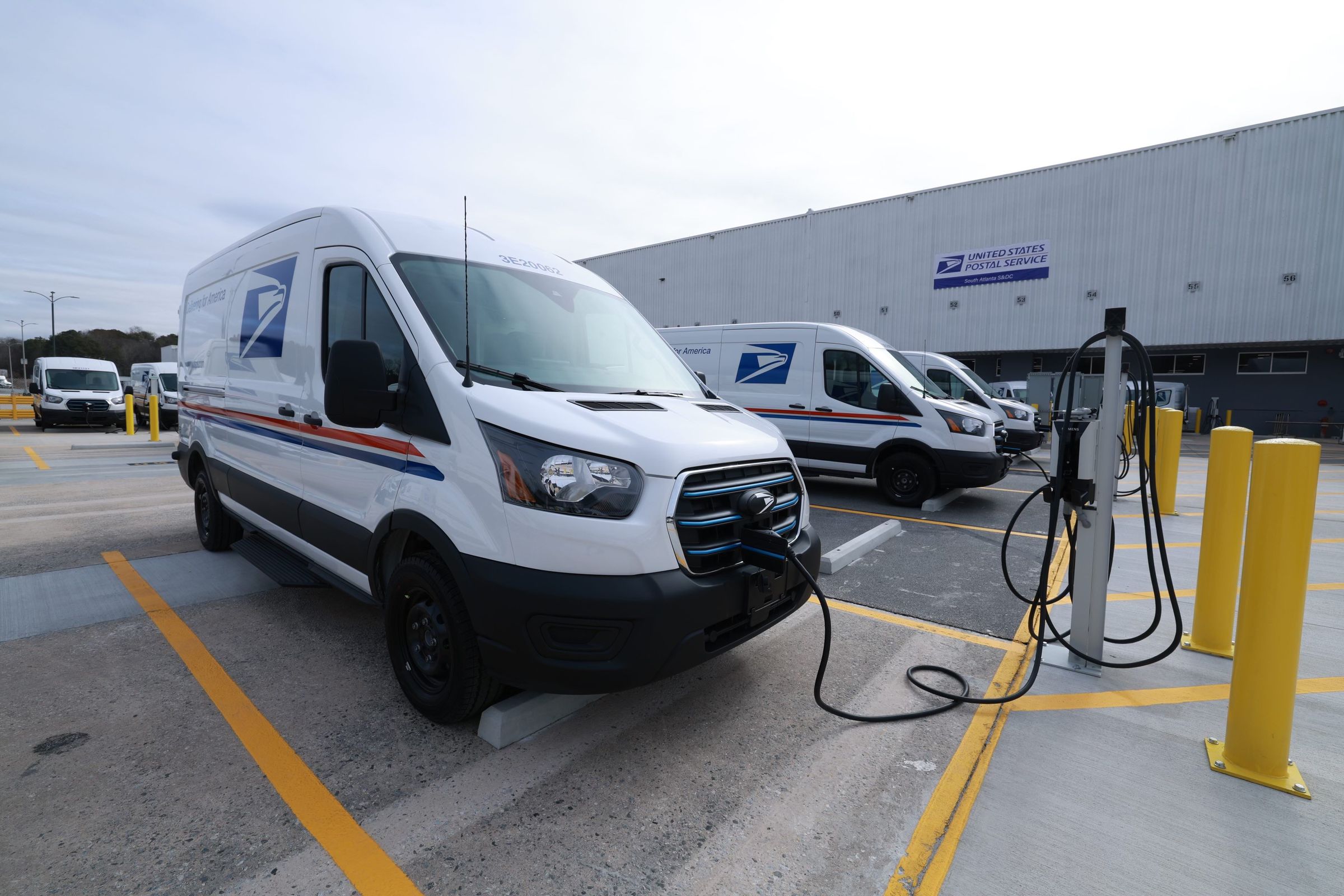 An electric delivery truck is plugged into a charging station in a parking lot.