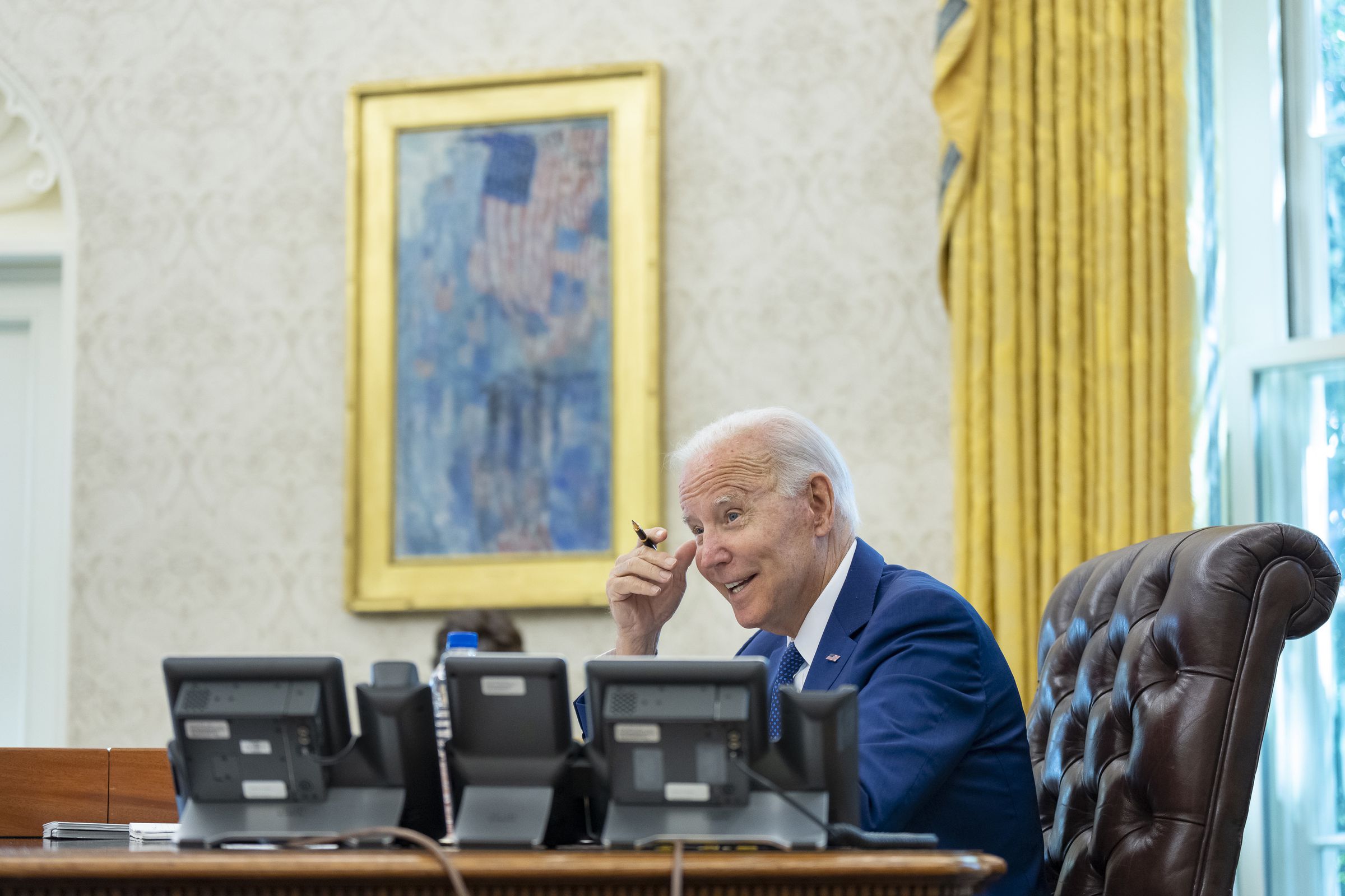 President Joe Biden sits smiling at a desk in the oval office, seen from his left behind a bank of phones.
