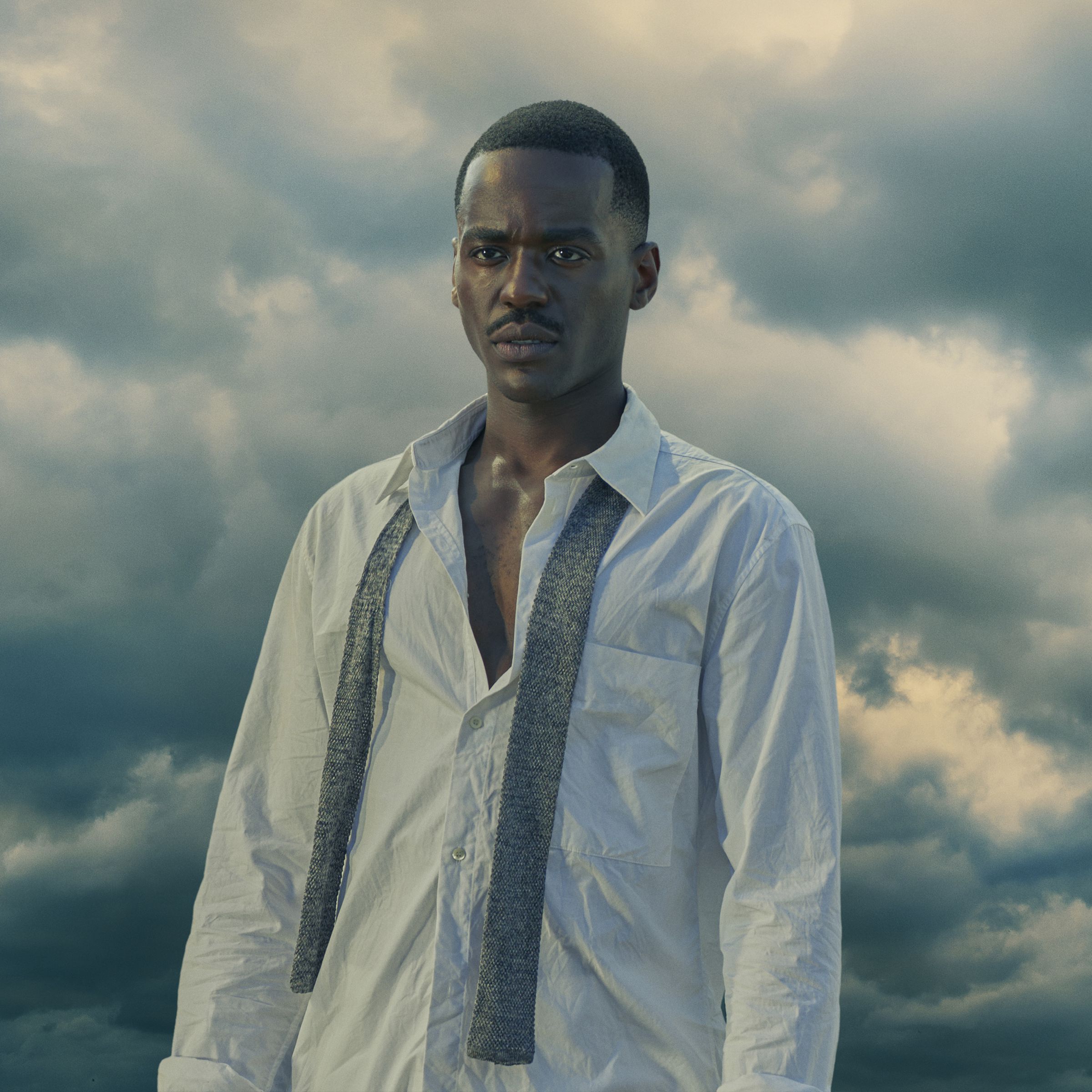 A Black man with a very close fade standing in a white button down shirt with an undone tie around his neck.