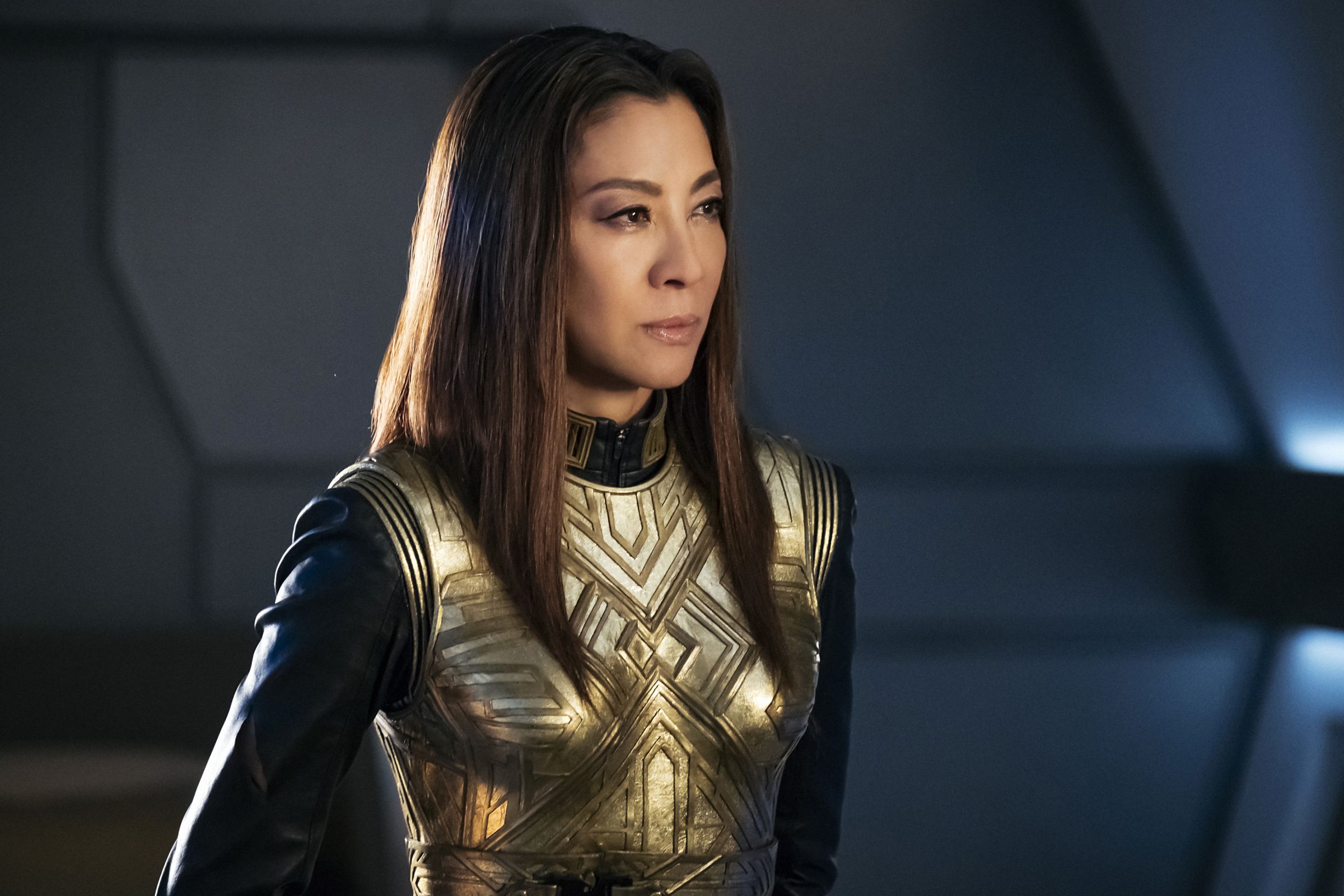 Michelle Yeoh stands straight, looking annoyed, and wearing a gold chest plate.