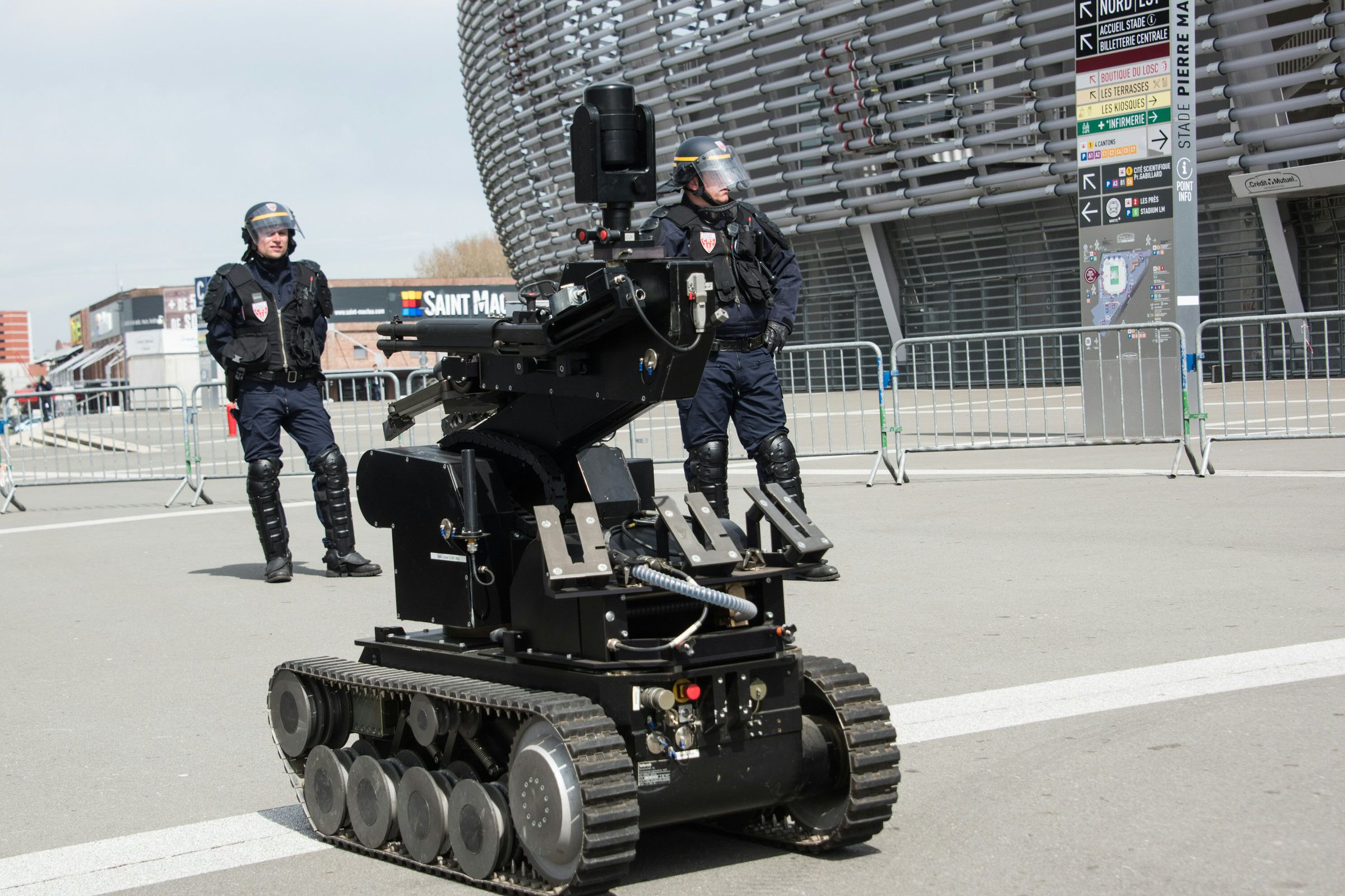 A picture of a black, tank-treaded robot outside a stadium flanked by two police officers in bomb protection gear.