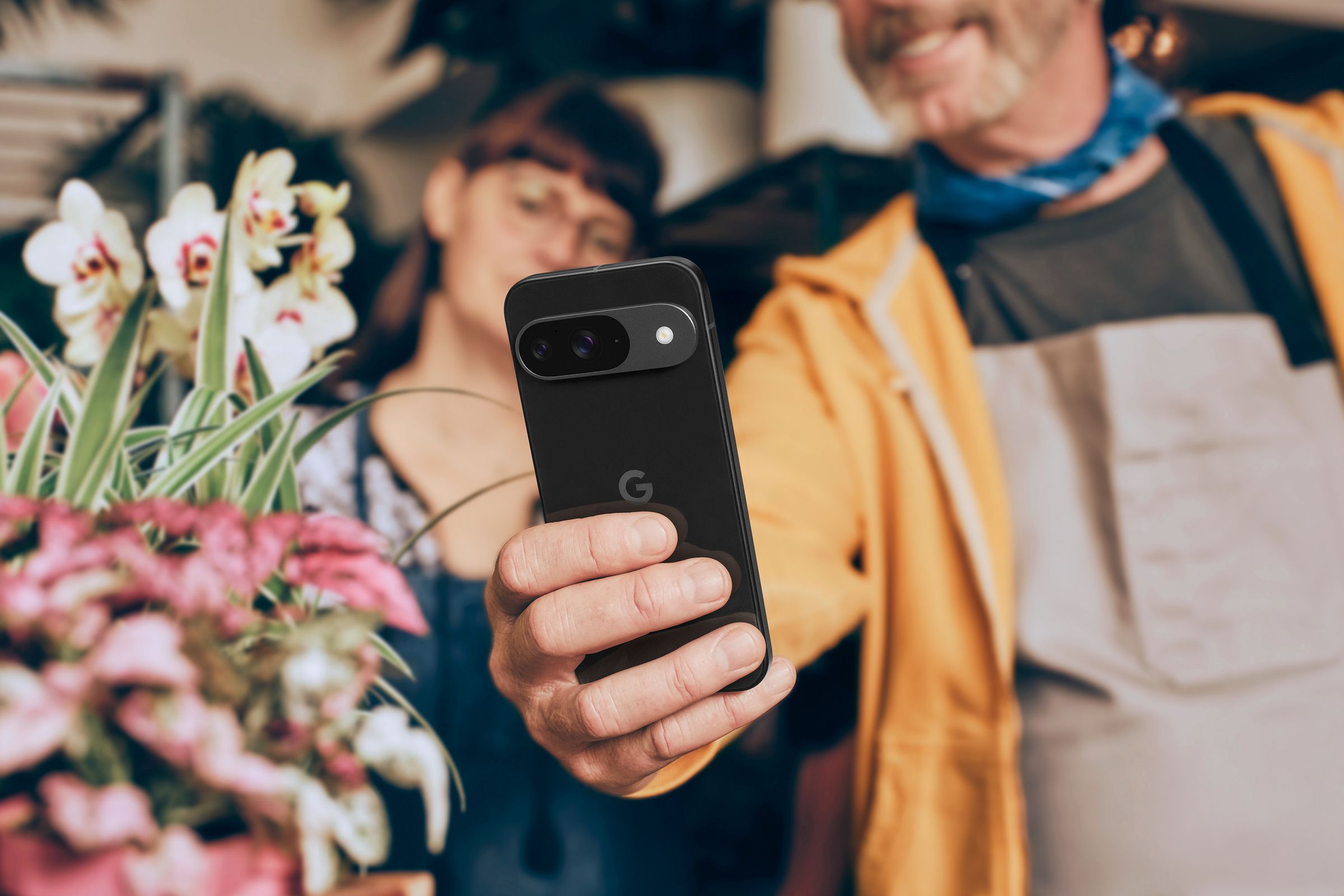 A person holding a Pixel 9 phone up at arm’s length to take a picture of flowers.